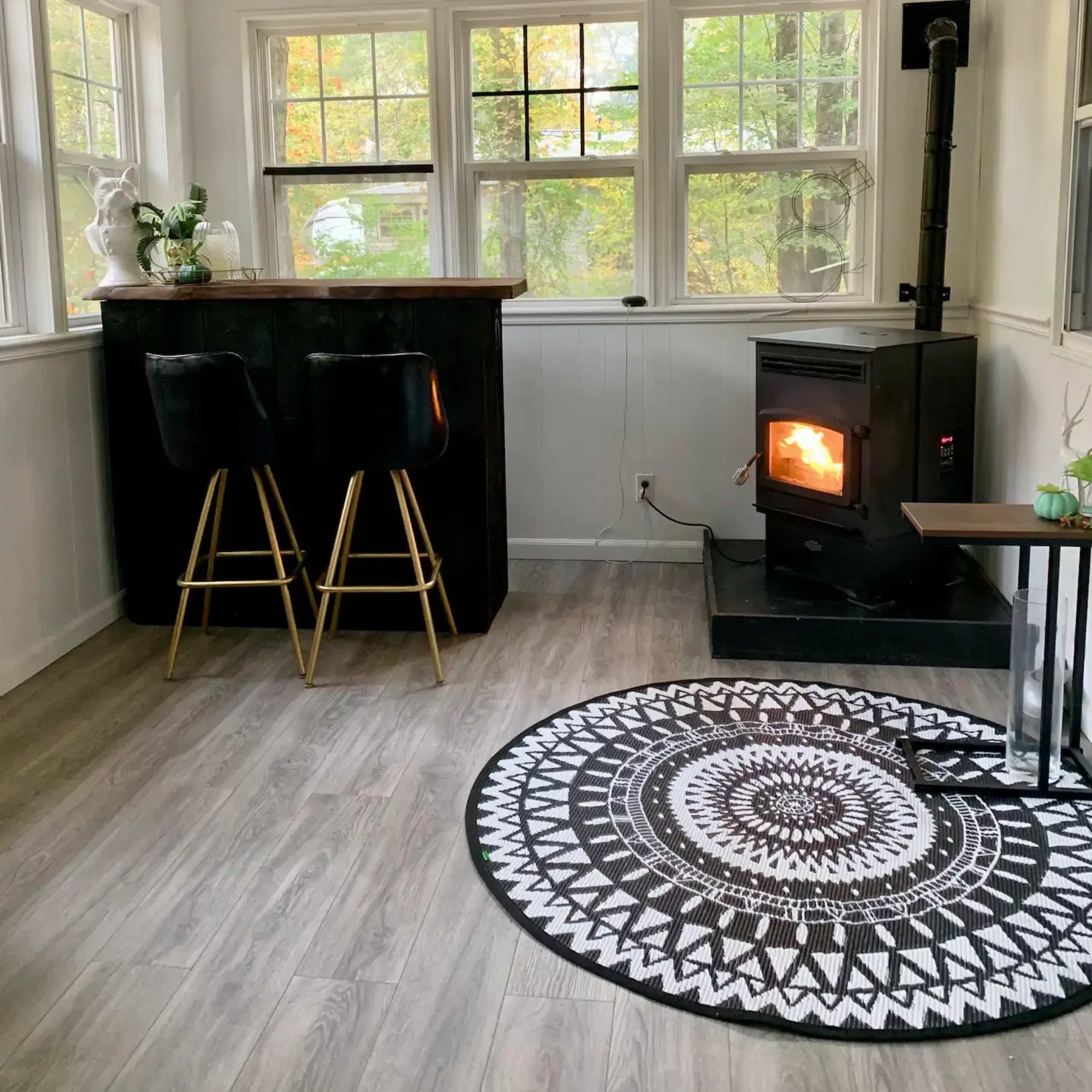 A cozy interior space with a black wood stove, a round patterned rug, and two high stools at a bar counter near large windows.