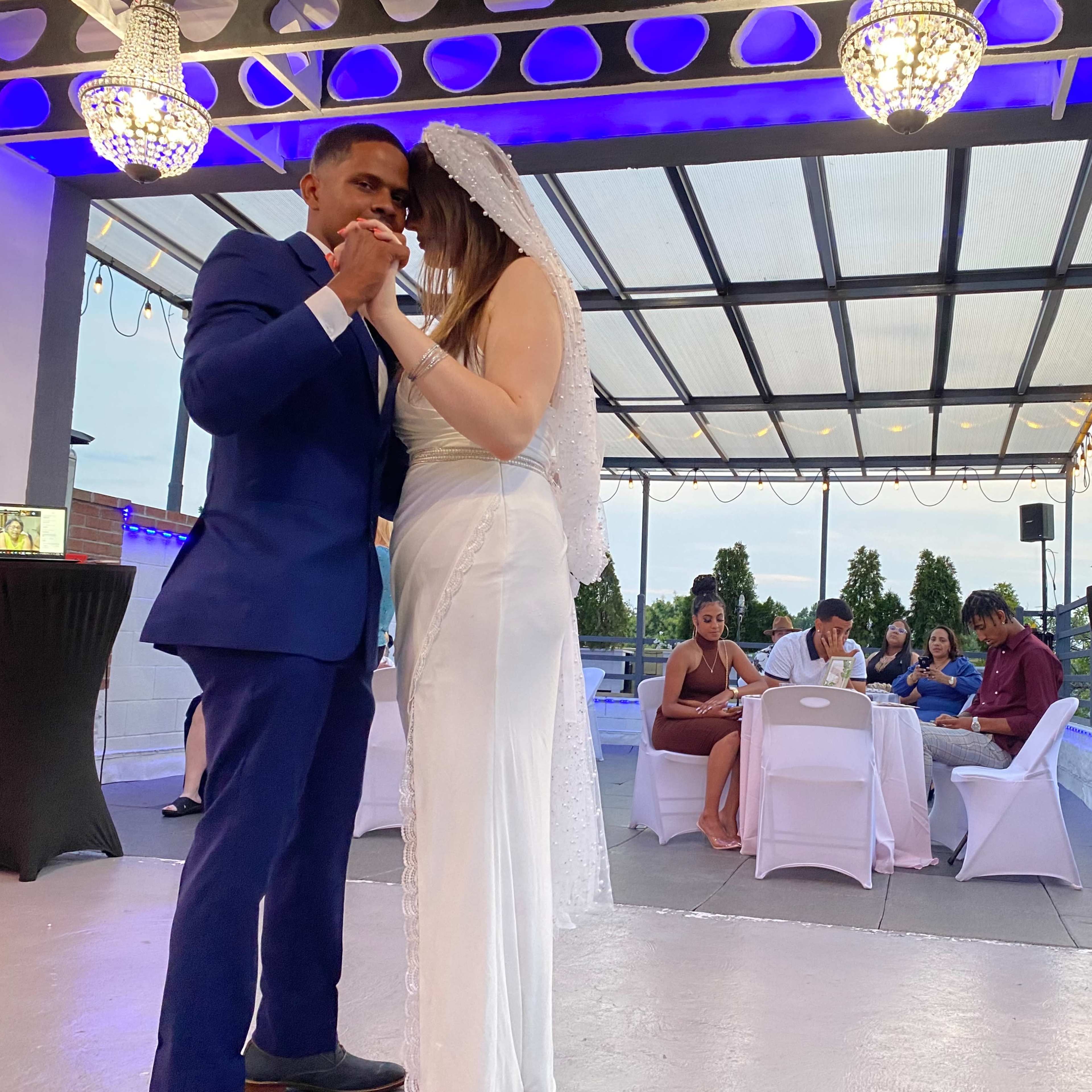 A couple dances together under hanging chandeliers at their wedding reception, while guests sit at tables in the background.