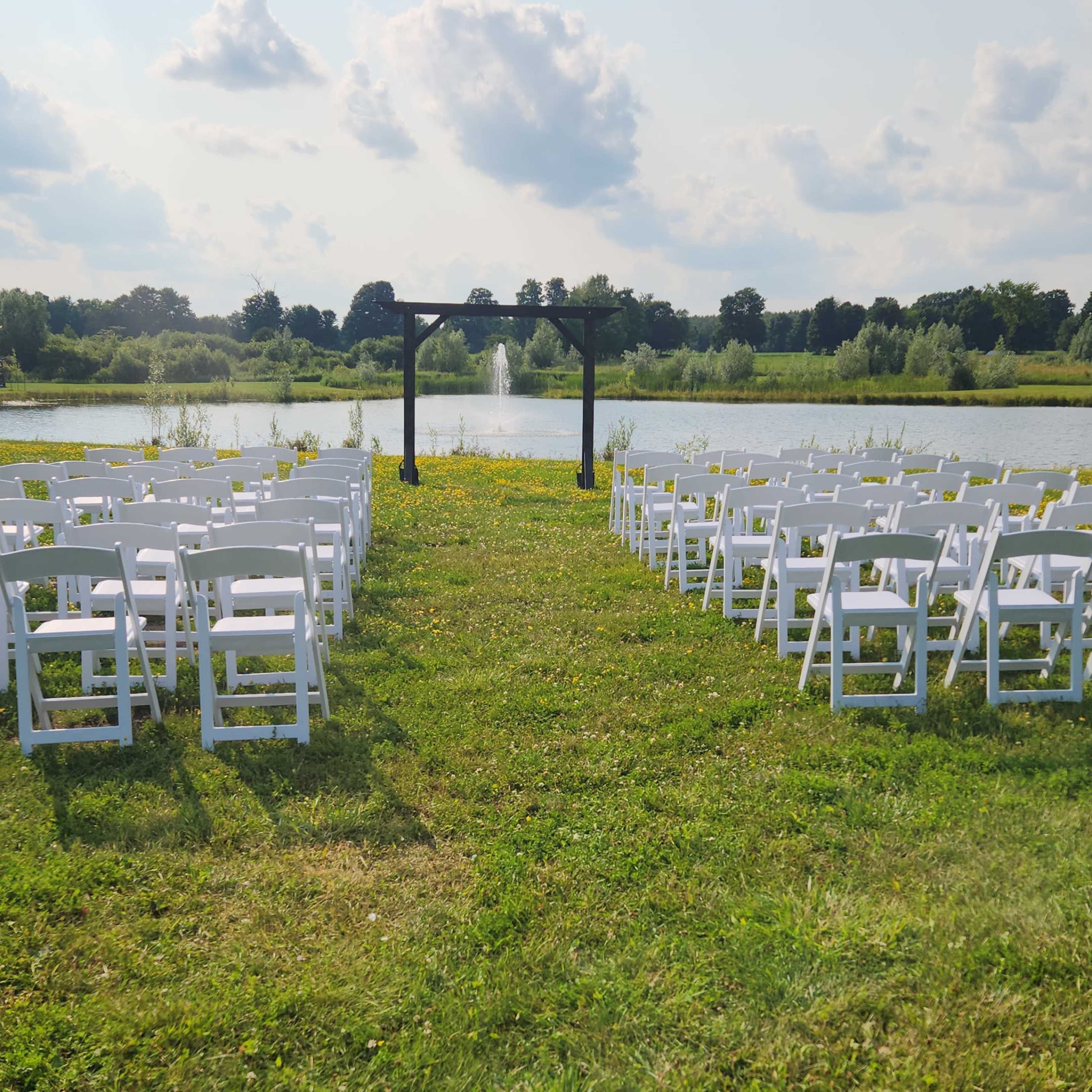 A row of white chairs is set up on a grassy area facing a wooden arch by a pond with a fountain in the background.