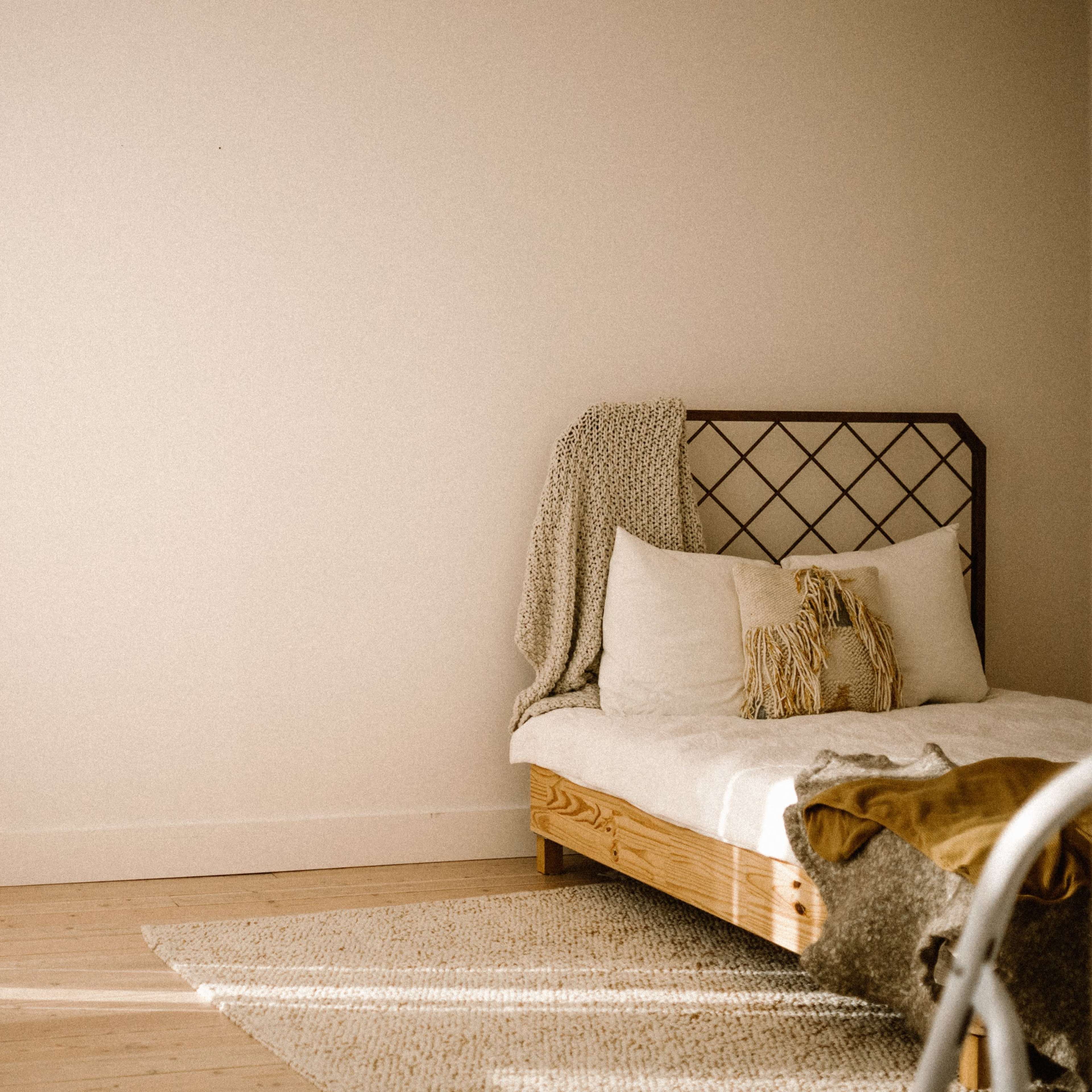 A minimalistic room features a light-colored bed with decorative pillows and a knitted throw, positioned next to a large window with sheer curtains.