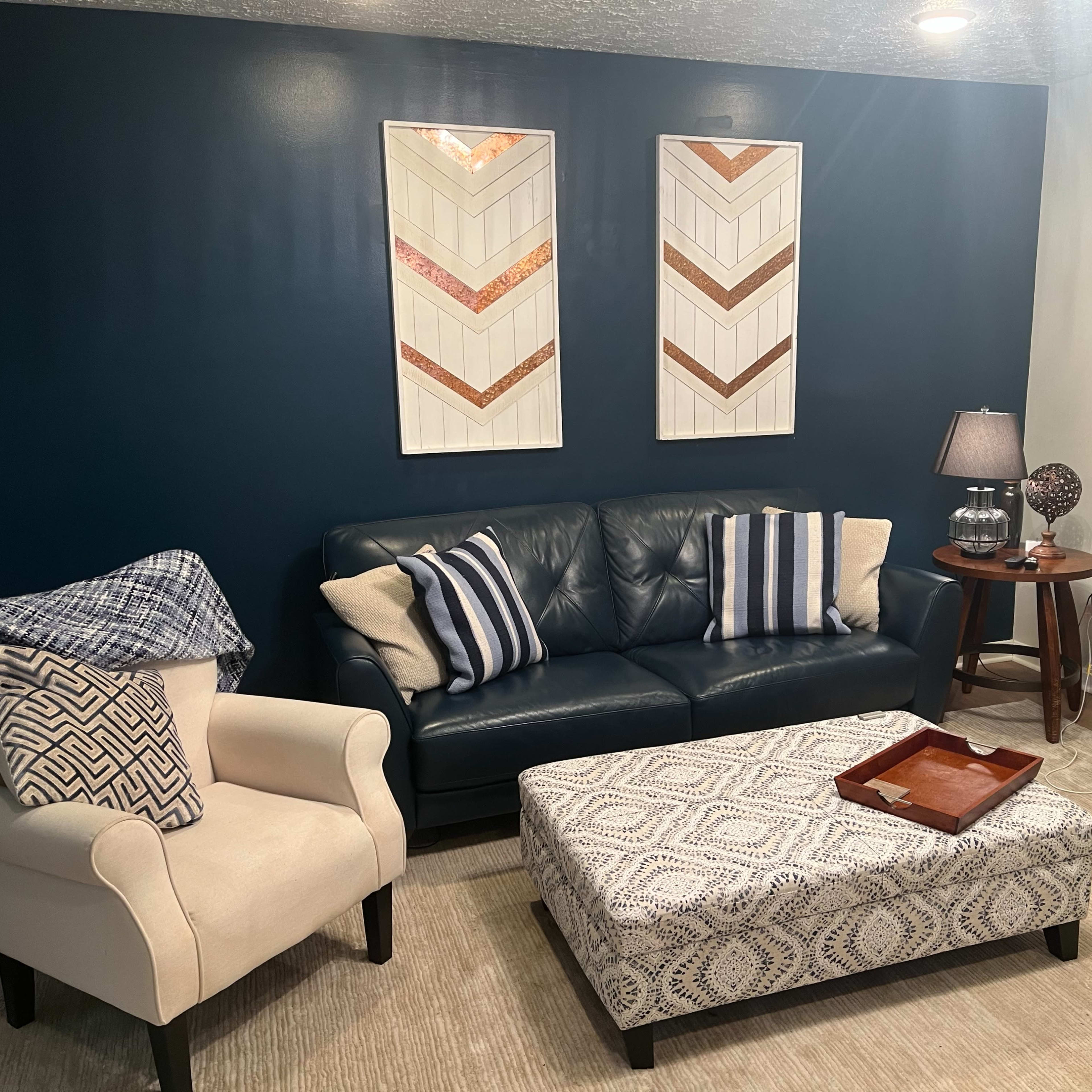 A living room with a dark blue accent wall, a navy sofa, two cream-colored armchairs, and a patterned ottoman in the center of the seating area.