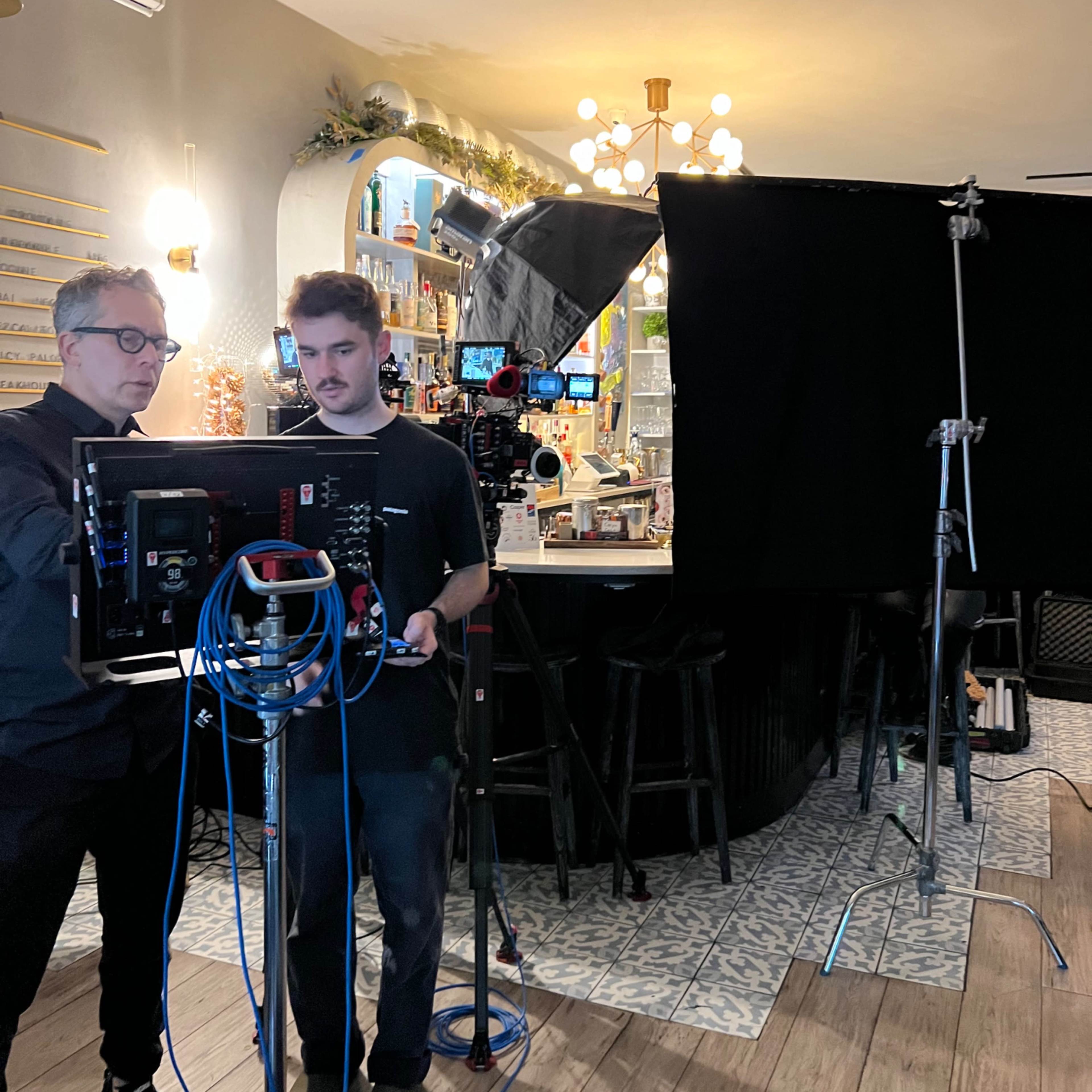 Two men are setting up a camera in a bar, with lighting equipment and a large black backdrop in the background.