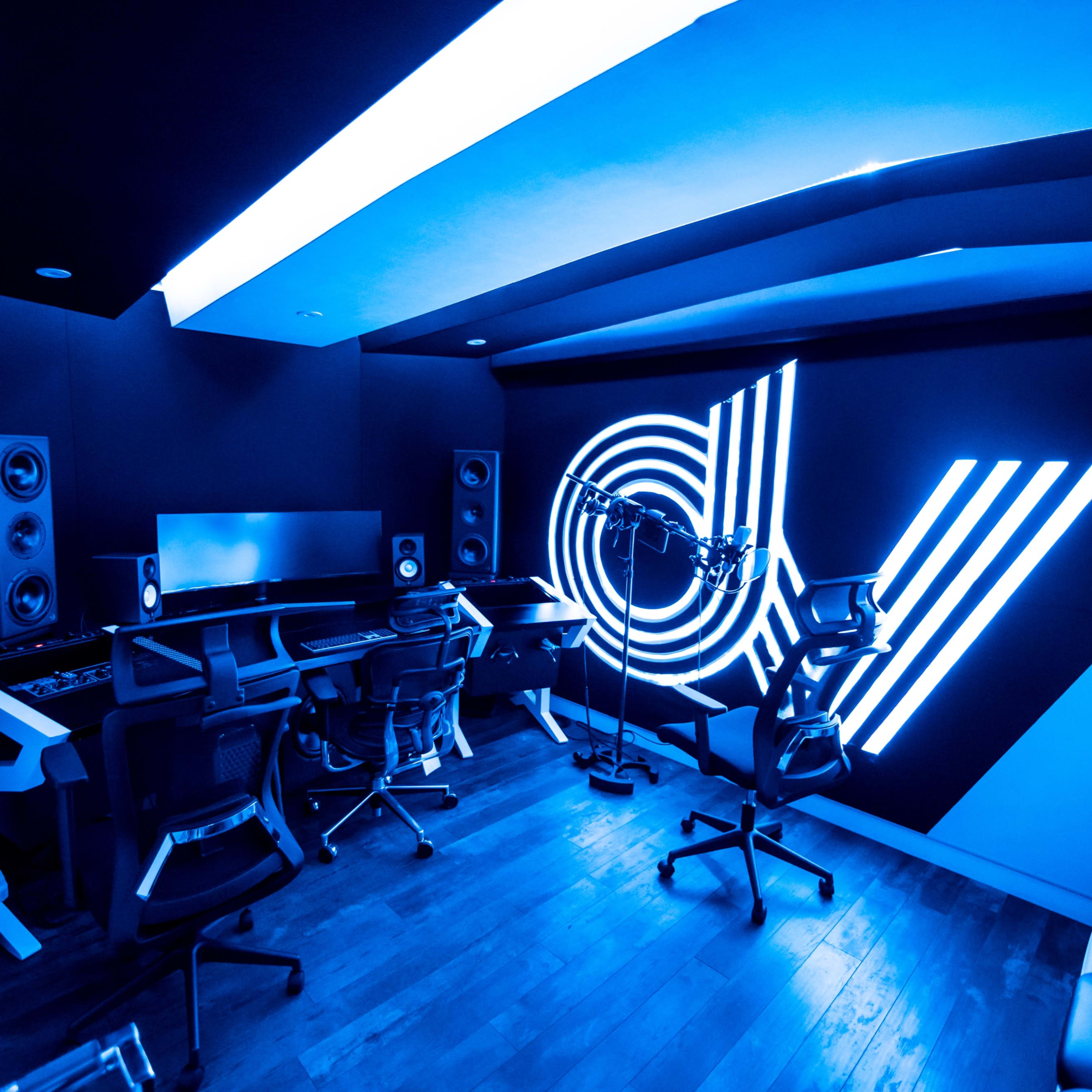 A modern music studio features blue lighting, sleek workstations, and sound equipment arranged against a black wall.