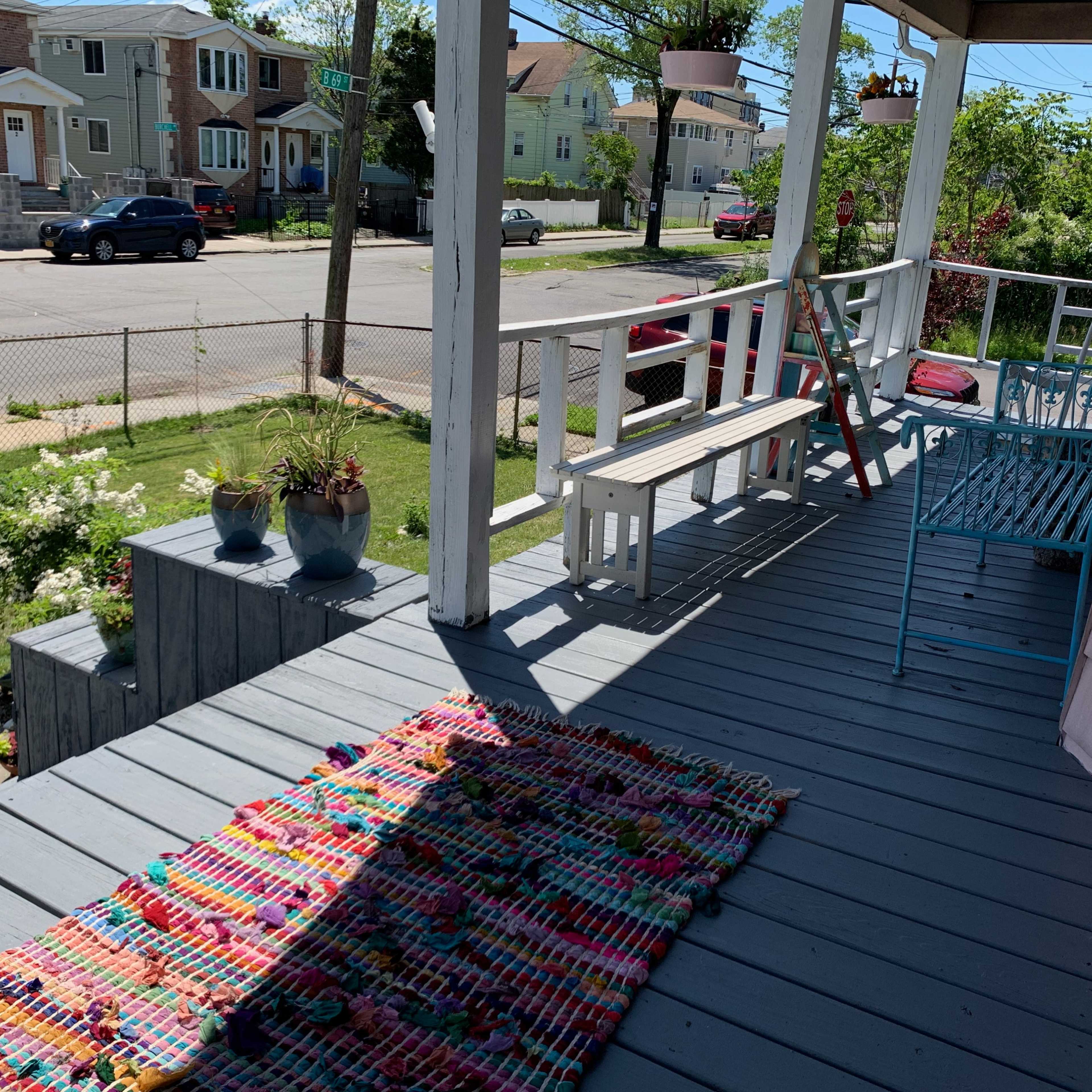 A porch with a colorful rug, potted plants, and a blue chair, overlooking a street with houses in the background.