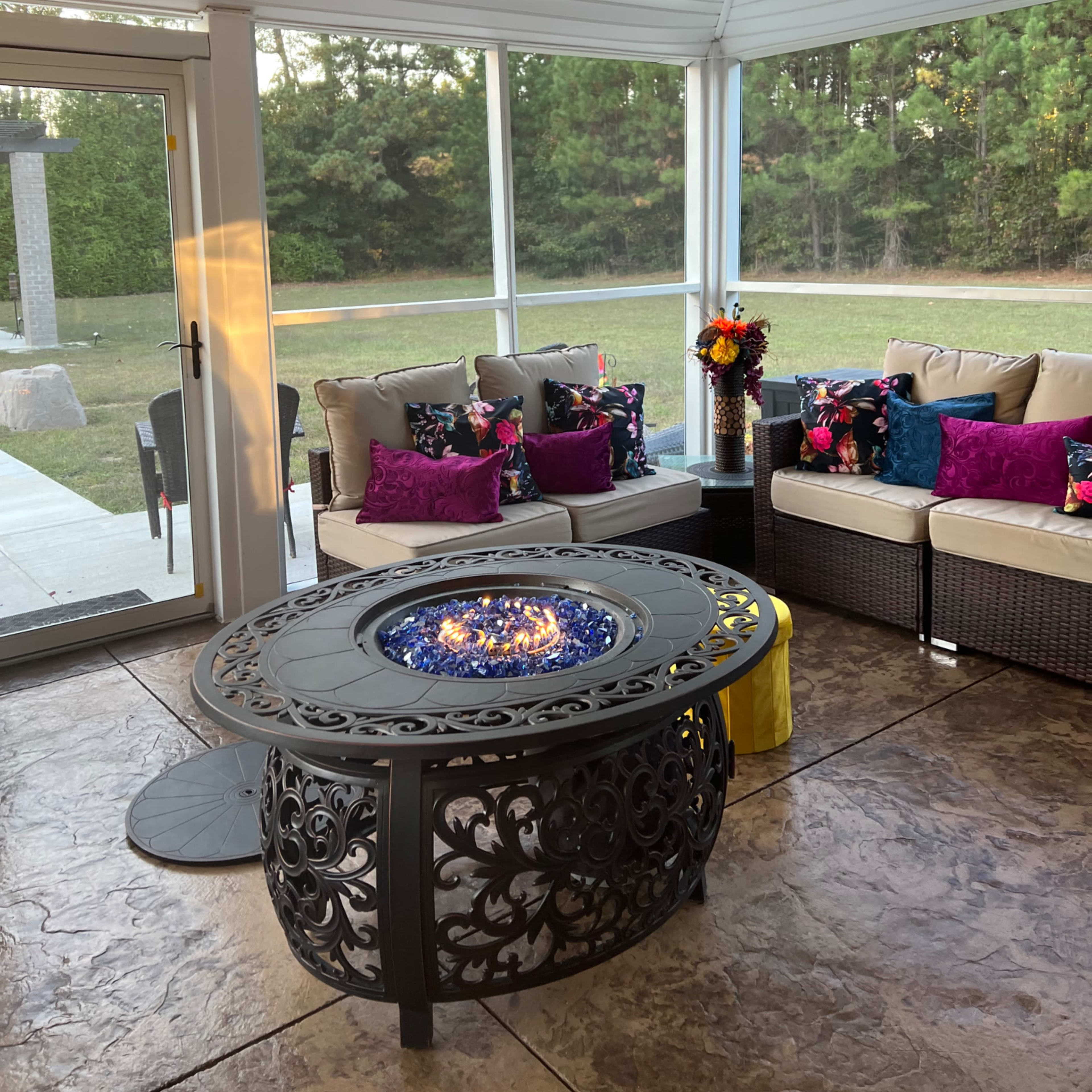 A screened porch with a decorative fire table in the center, surrounded by a sectional sofa adorned with colorful cushions and a view of a grassy area beyond.