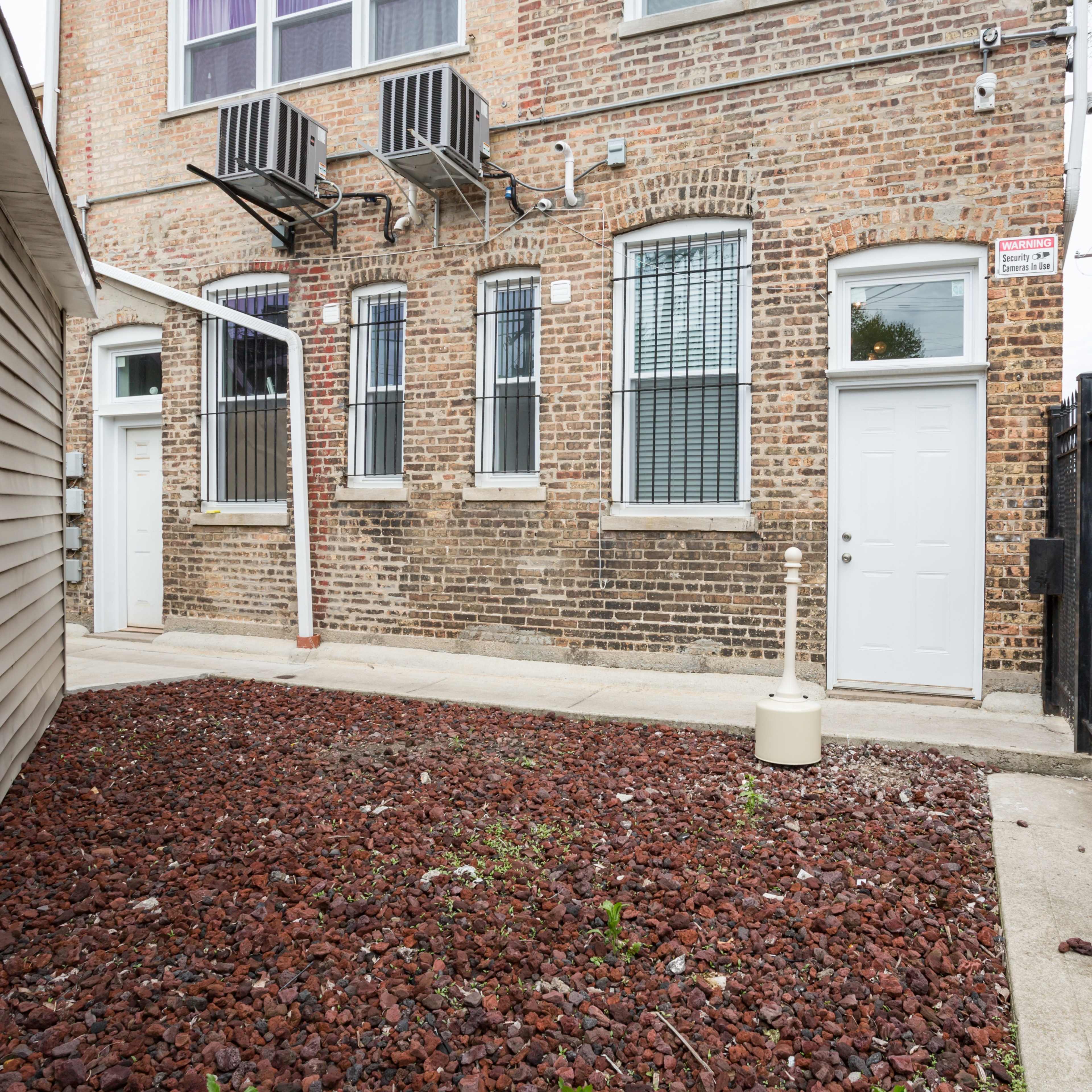 A narrow outdoor area with a gravel surface, flanked by a brick building and a wooden structure, featuring doors and windows.