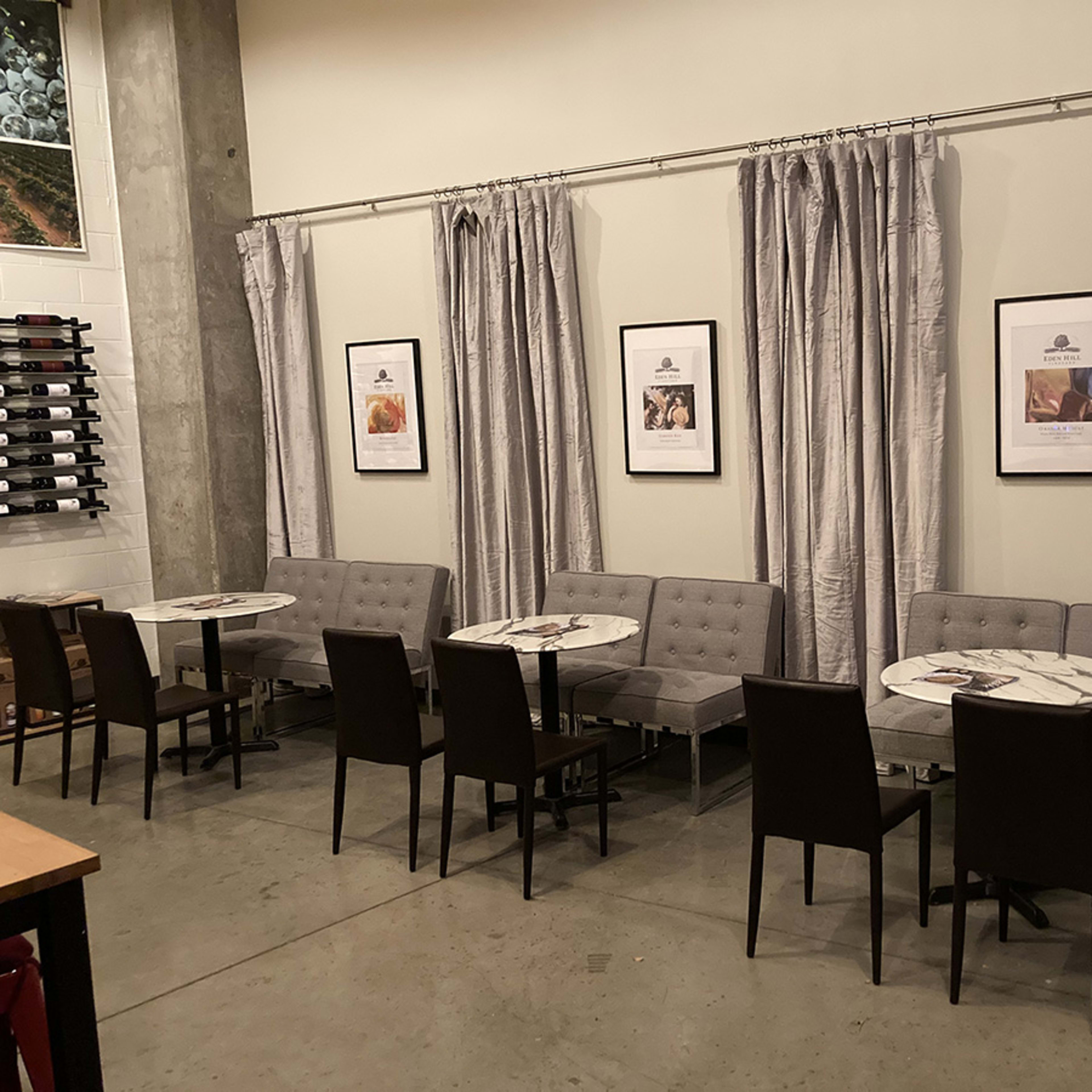A restaurant interior with several gray benches and tables arranged against a wall, accompanied by framed pictures and floor-to-ceiling curtains.