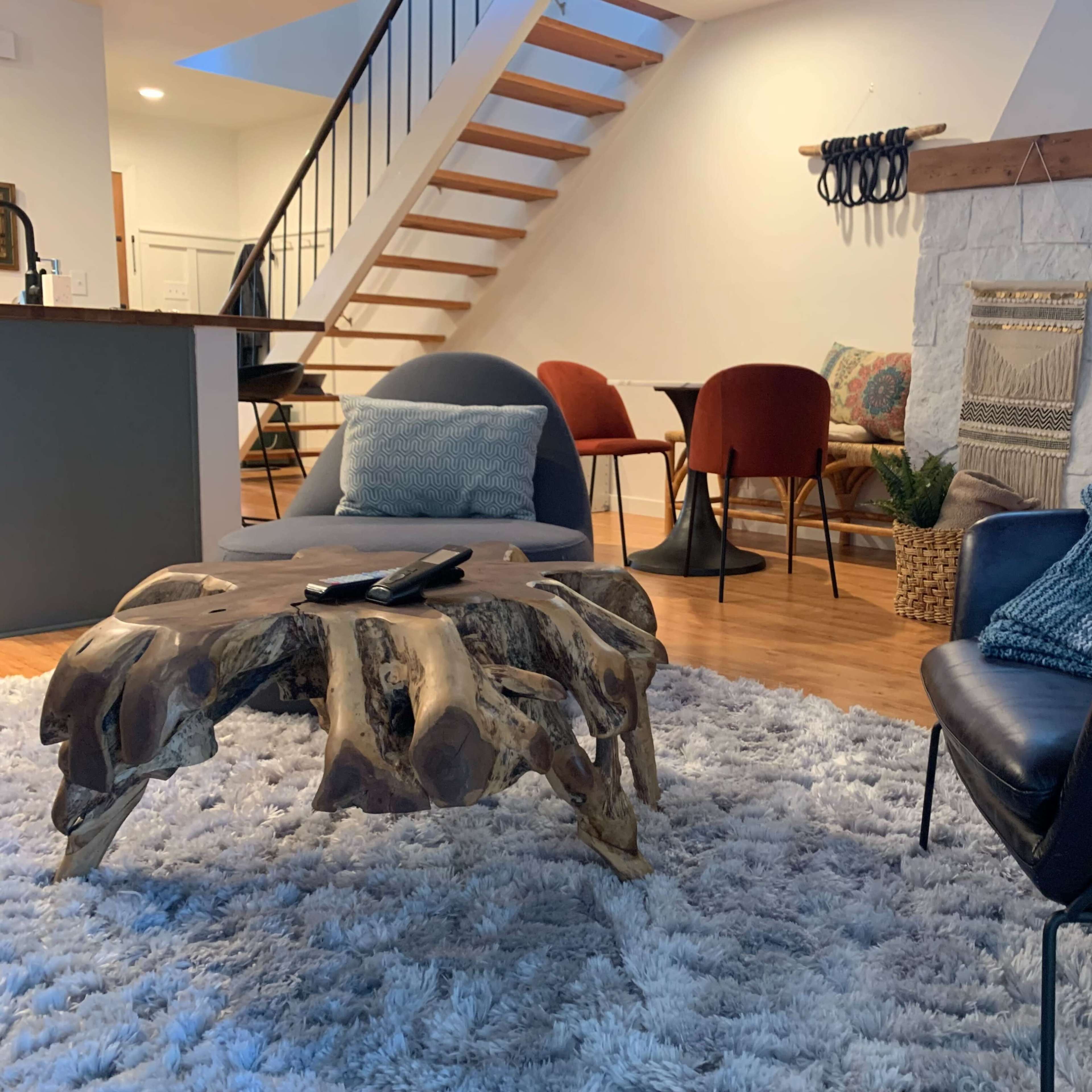 A modern living space featuring a wooden coffee table, two armchairs, a faux fur rug, and a staircase in the background.