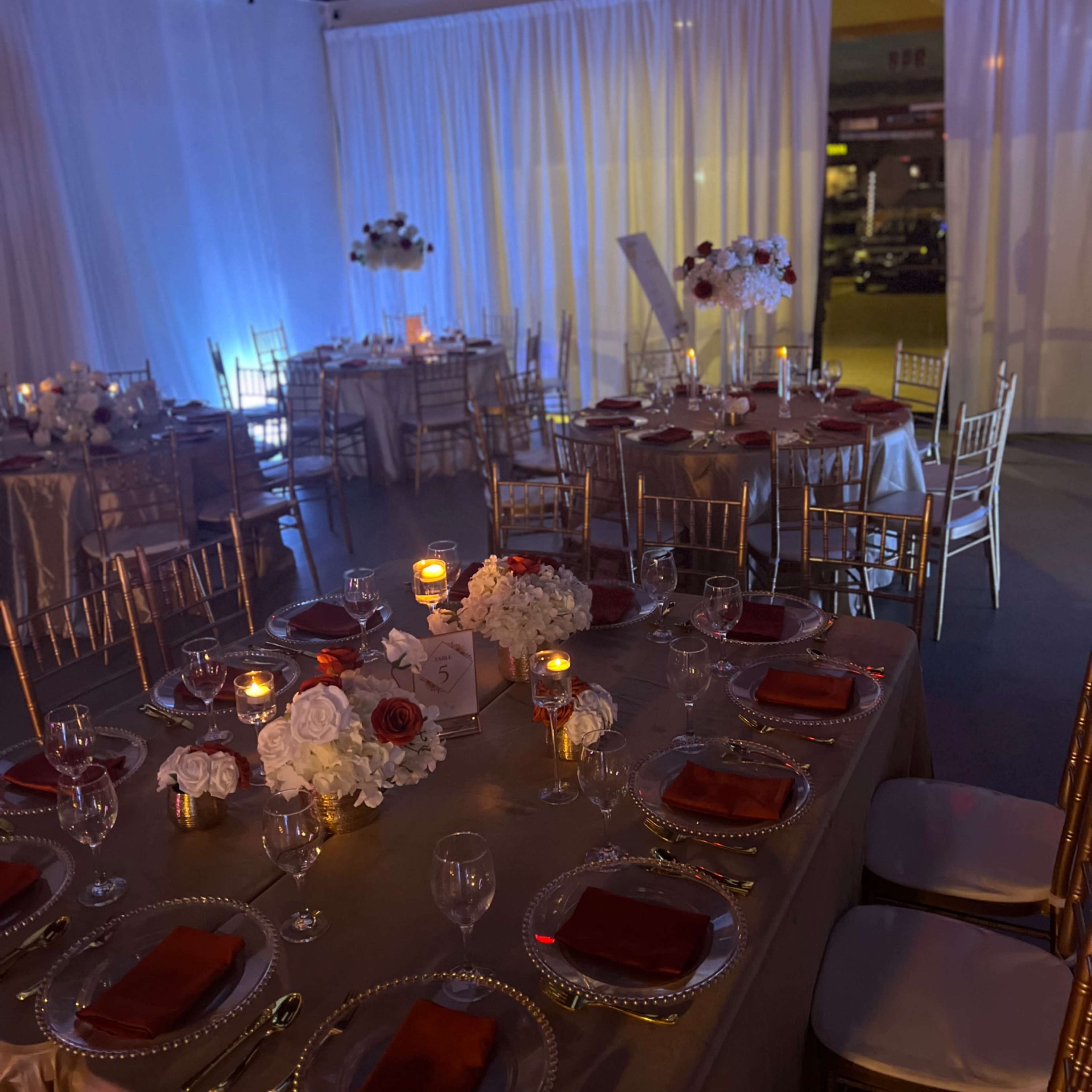 A banquet setup with elegantly arranged tables featuring gold chairs, white tablecloths, and centerpieces of flowers and candles, all under soft lighting.