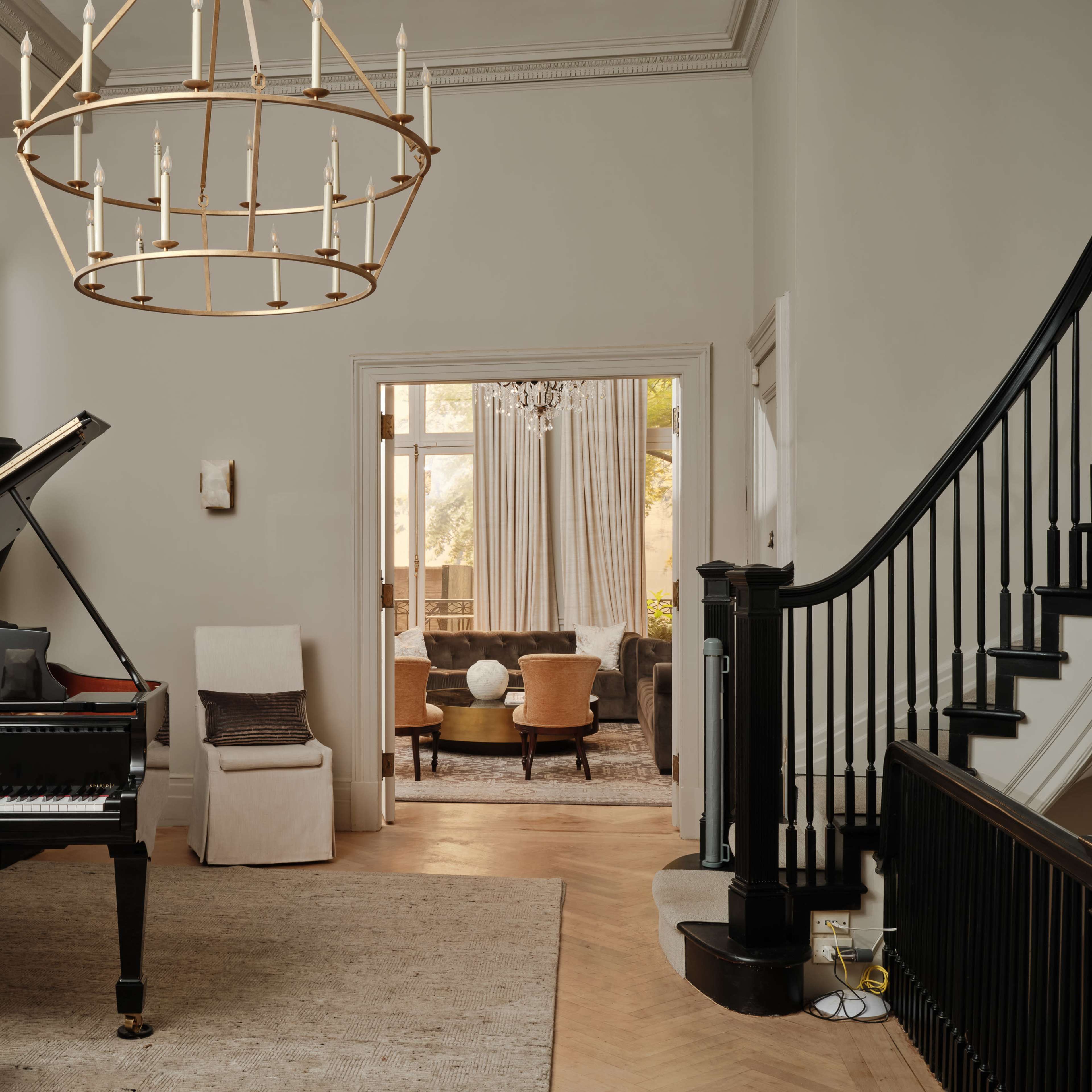 A grand piano with sheet music sits in a spacious entryway featuring a chandelier and a staircase, with an open living area visible through a doorway.