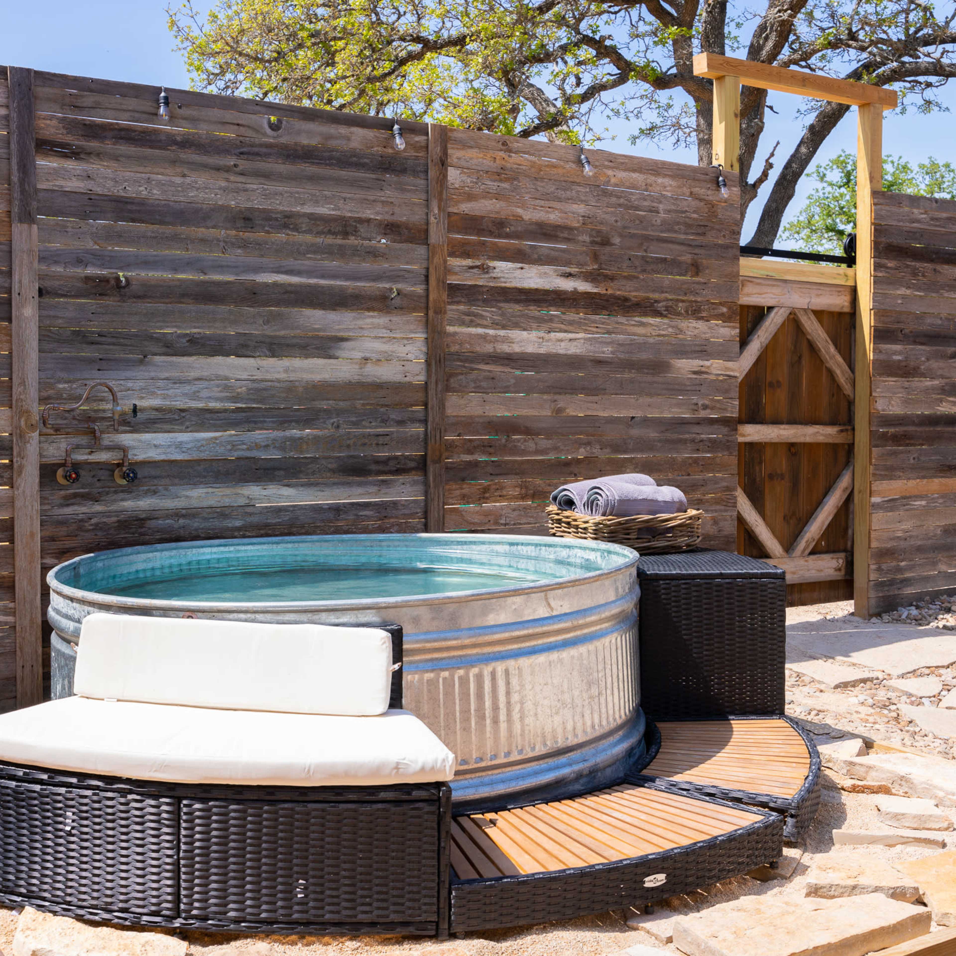 A round metal hot tub is set on a stone patio next to a wooden privacy fence, accompanied by a small seating area and a towel.