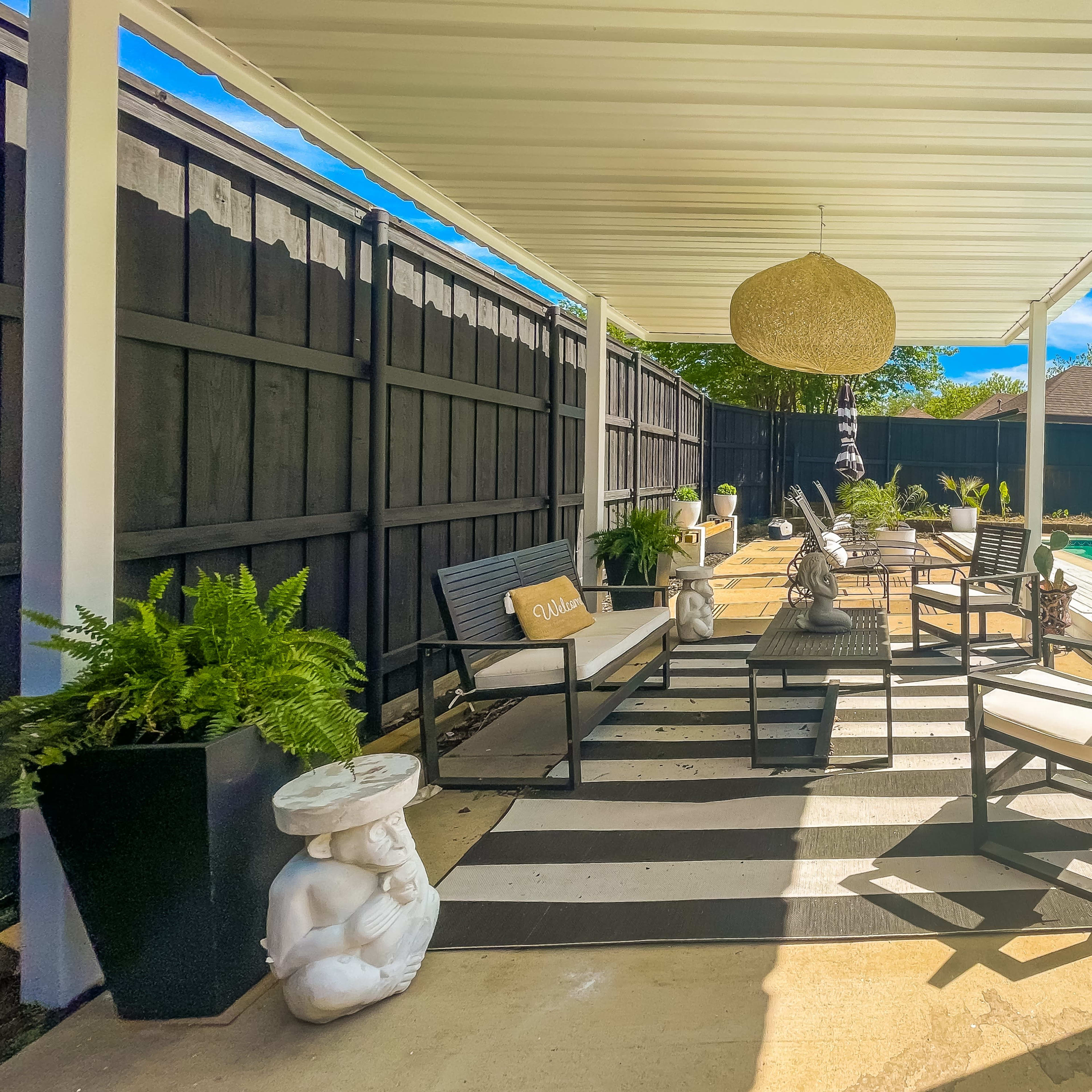 A covered outdoor seating area features black and white striped flooring, comfortable chairs, potted ferns, and decorative sculptures beside a pool.