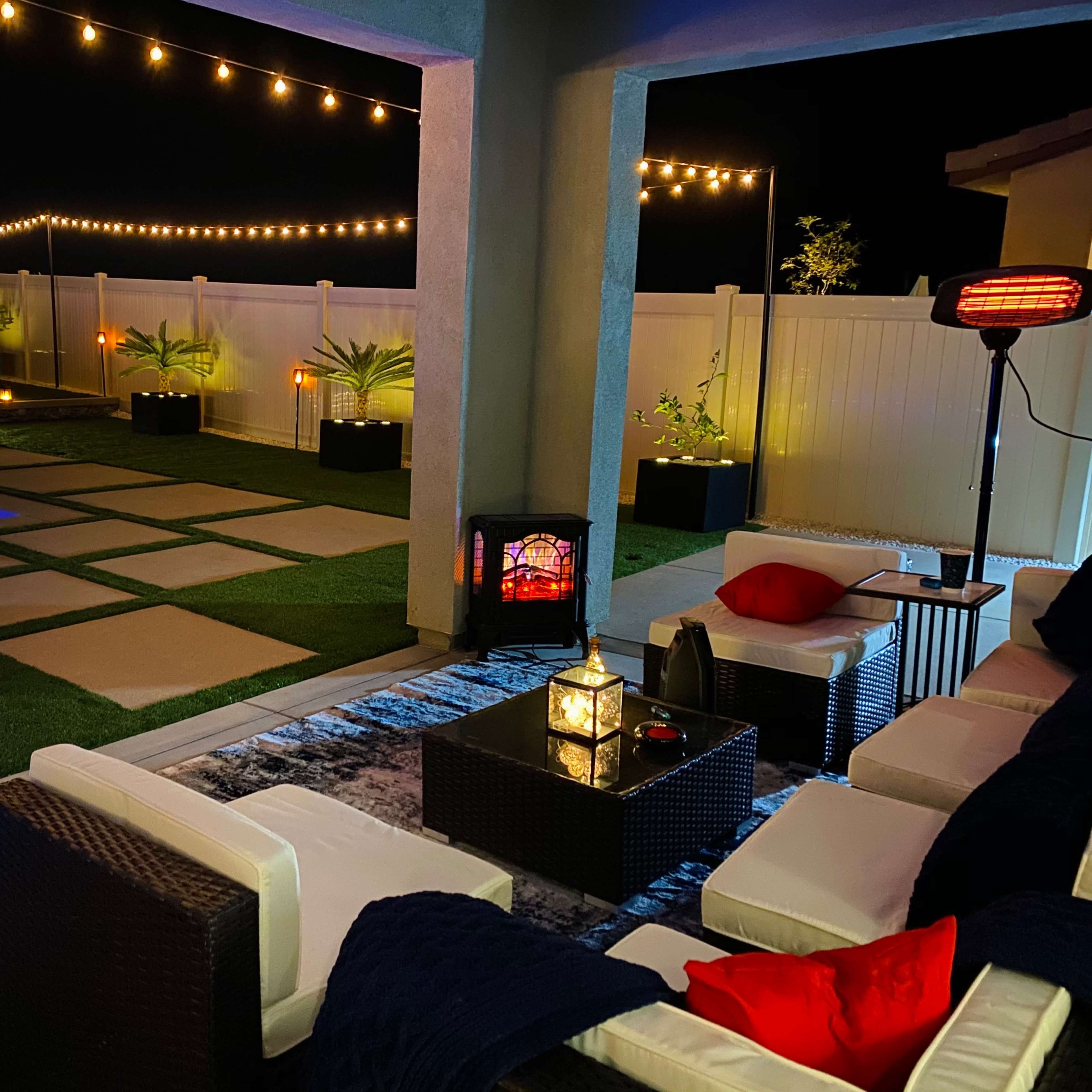 A cozy outdoor seating area at night, featuring a heated lamp, string lights, and a swimming pool with blue lighting.