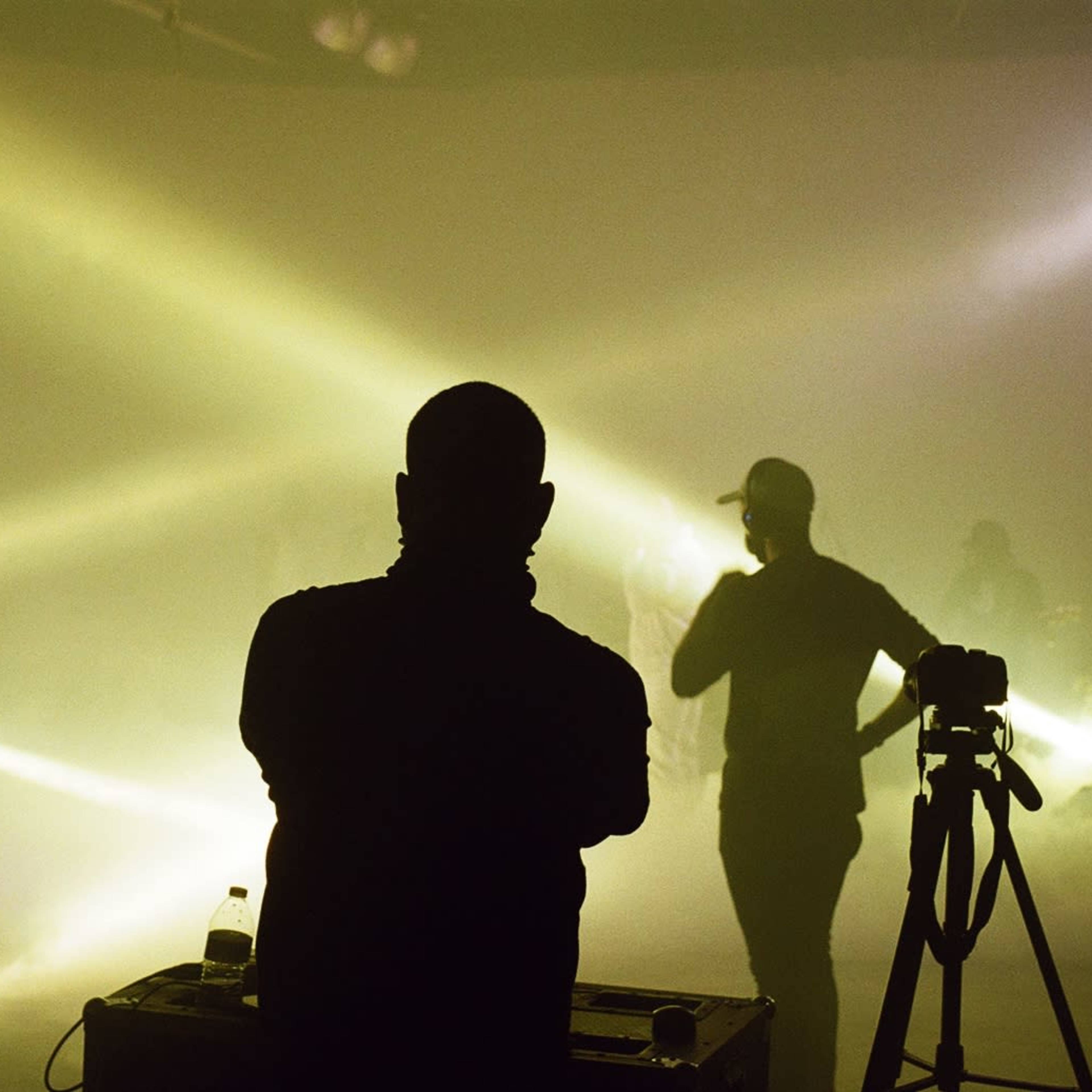 A silhouetted figure stands in front of stage lights while others perform in the background.