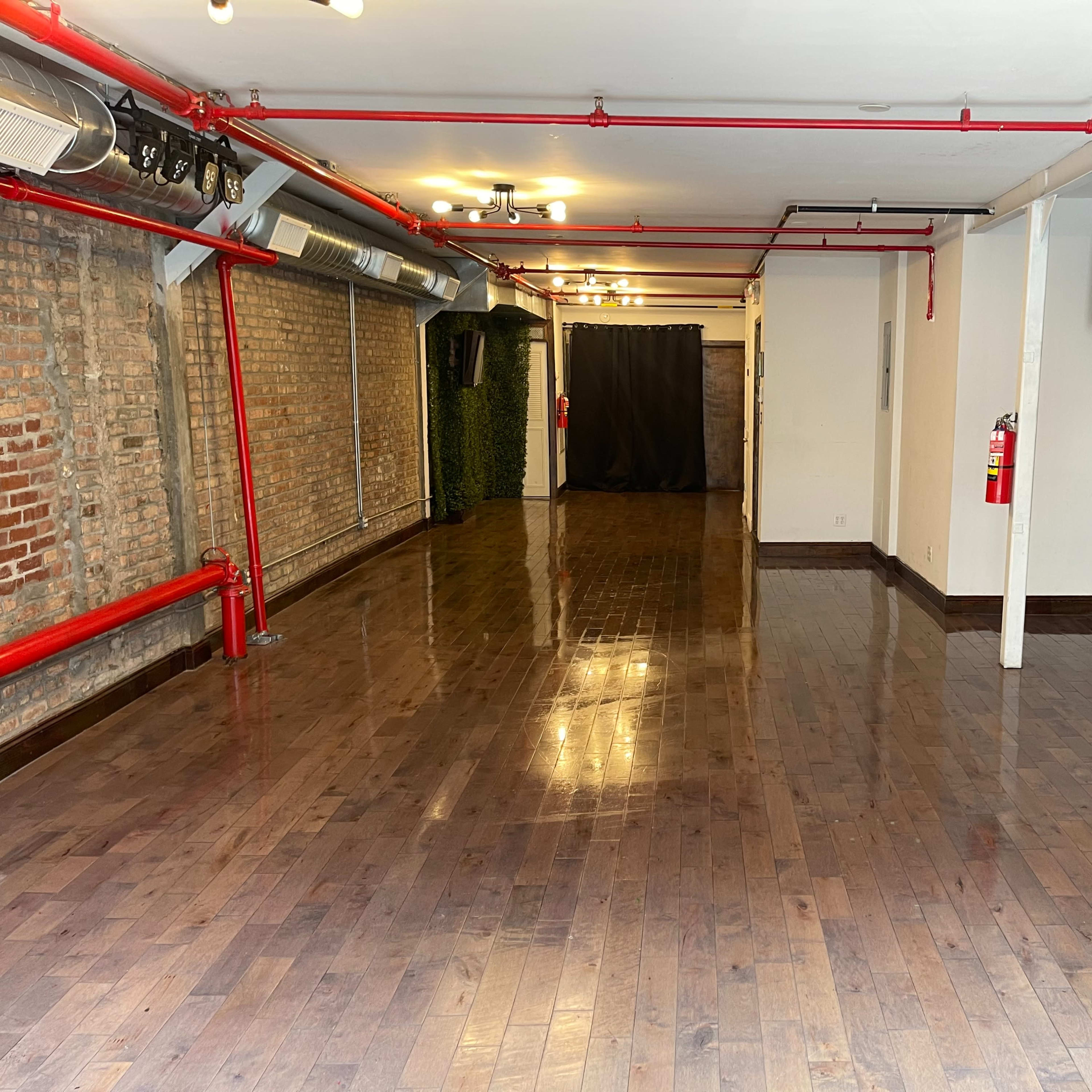 An empty room with polished wooden floors, exposed brick walls, and red pipes along the ceiling.
