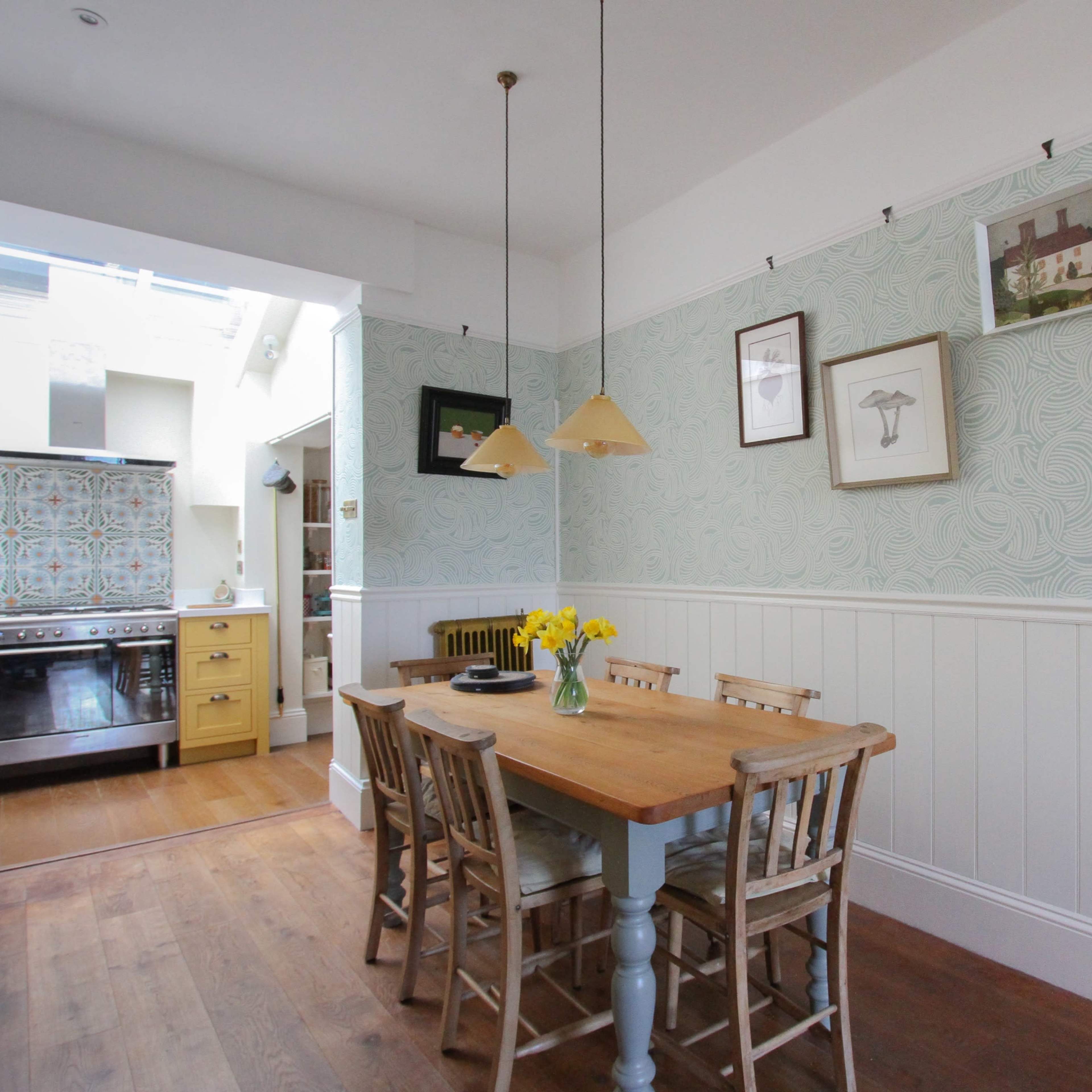 A dining area features a wooden table with chairs, yellow flowers in a vase, and patterned wallpaper, leading into a kitchen space with a stove and colorful tile backsplash.