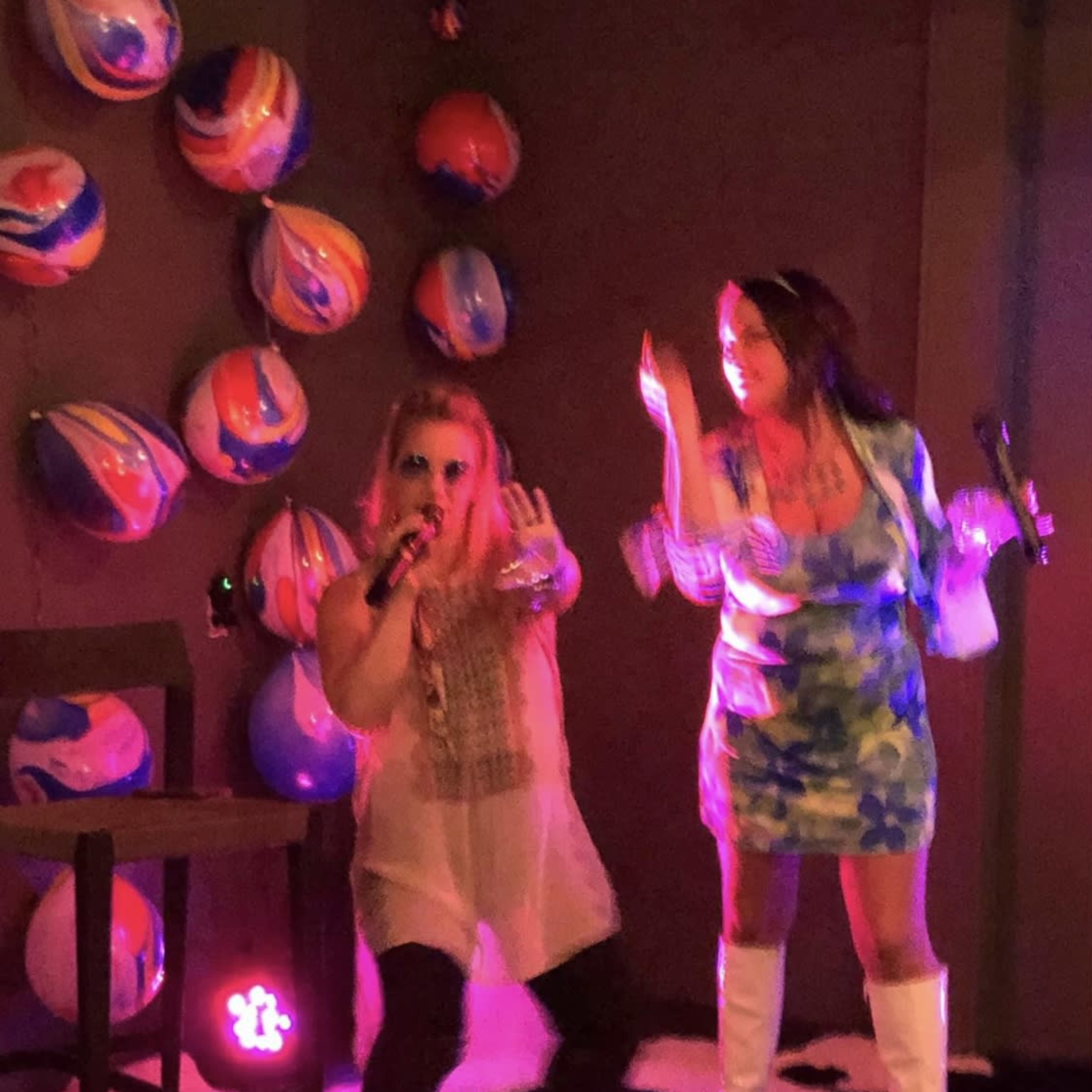Two women are performing karaoke with microphones on a stage decorated with colorful balls and a spotlight.