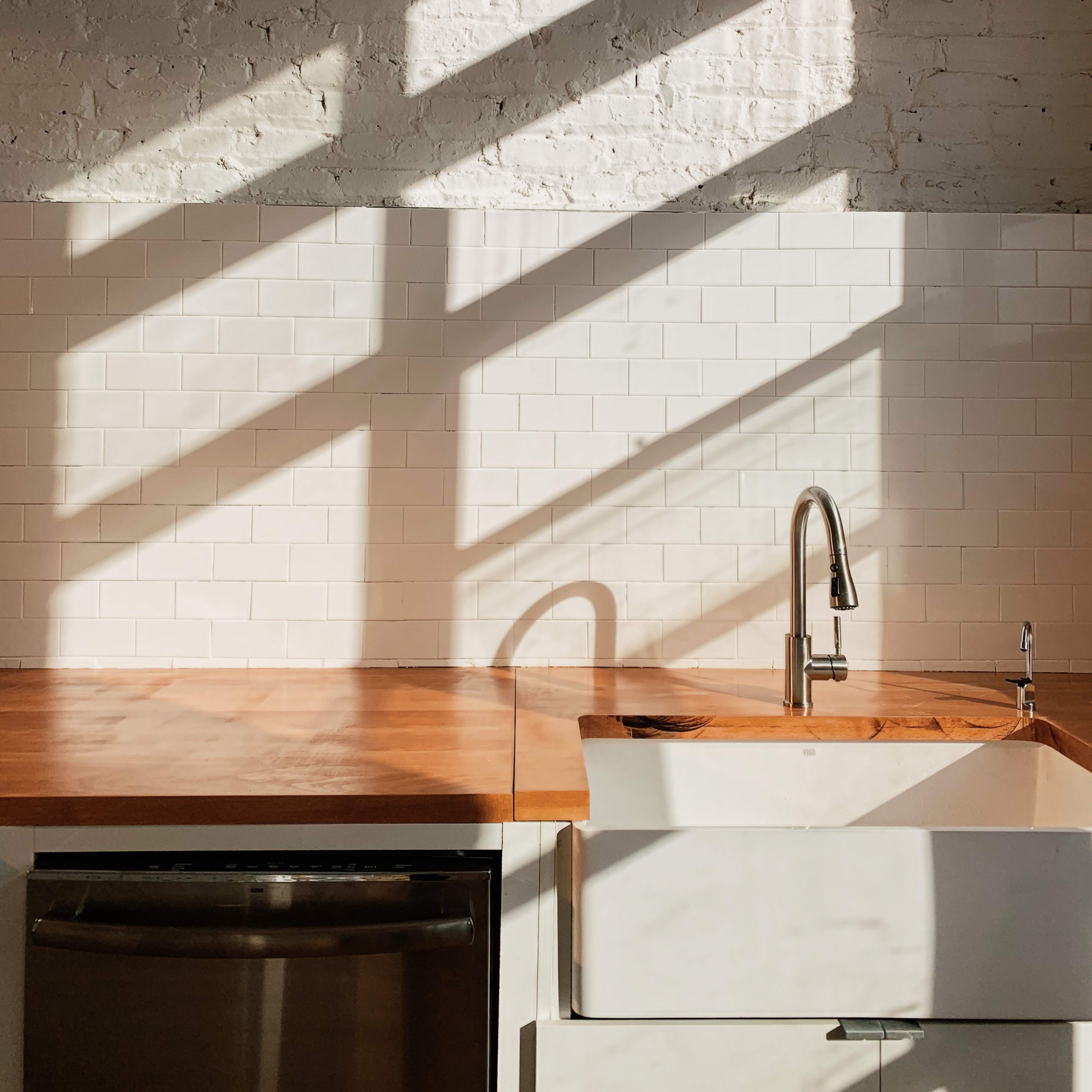 A kitchen sink area with a wooden countertop and white subway tile backsplash, illuminated by sunlight casting shadows across the surfaces.