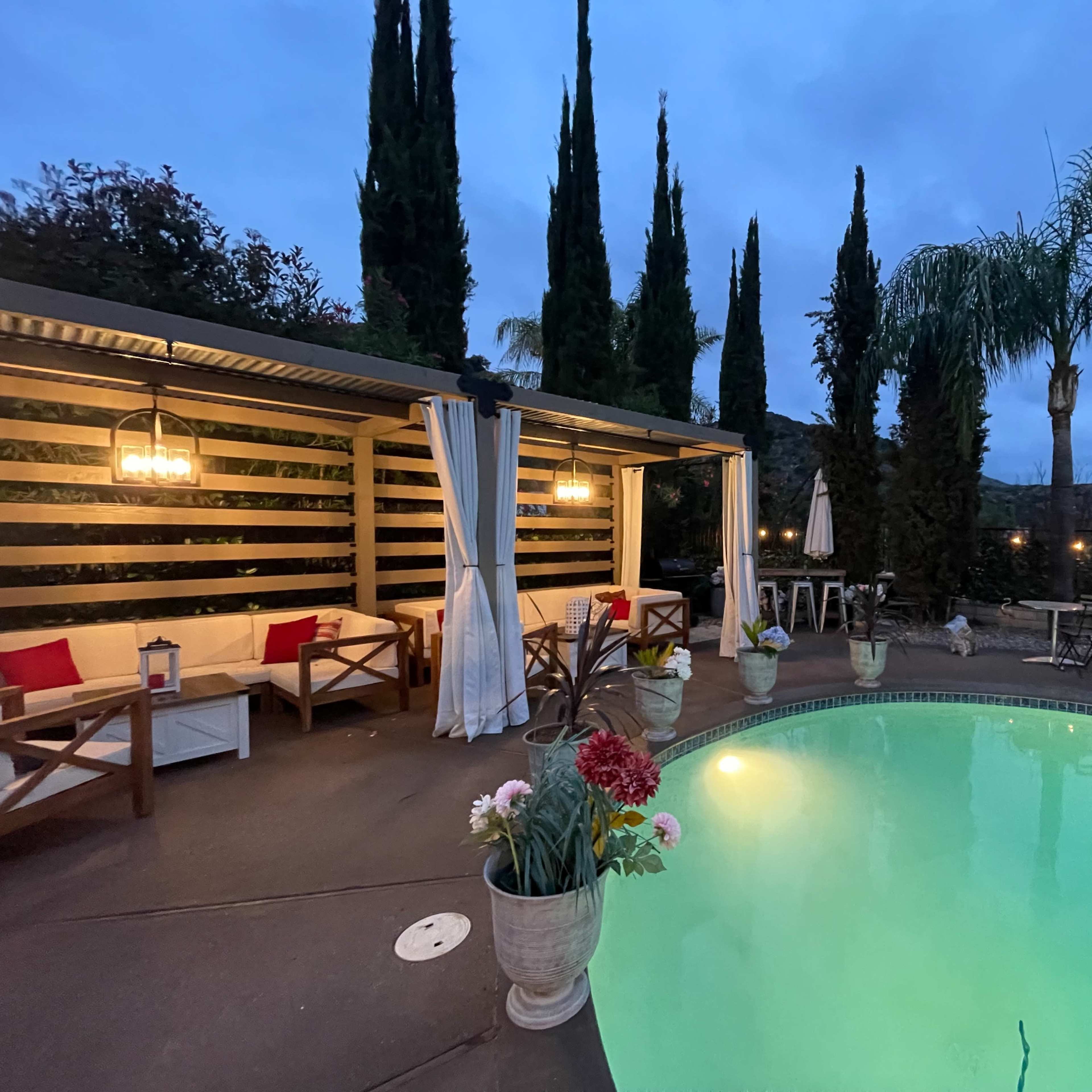 A swimming pool surrounded by tall greenery and a shaded seating area with lanterns, set against a twilight sky.