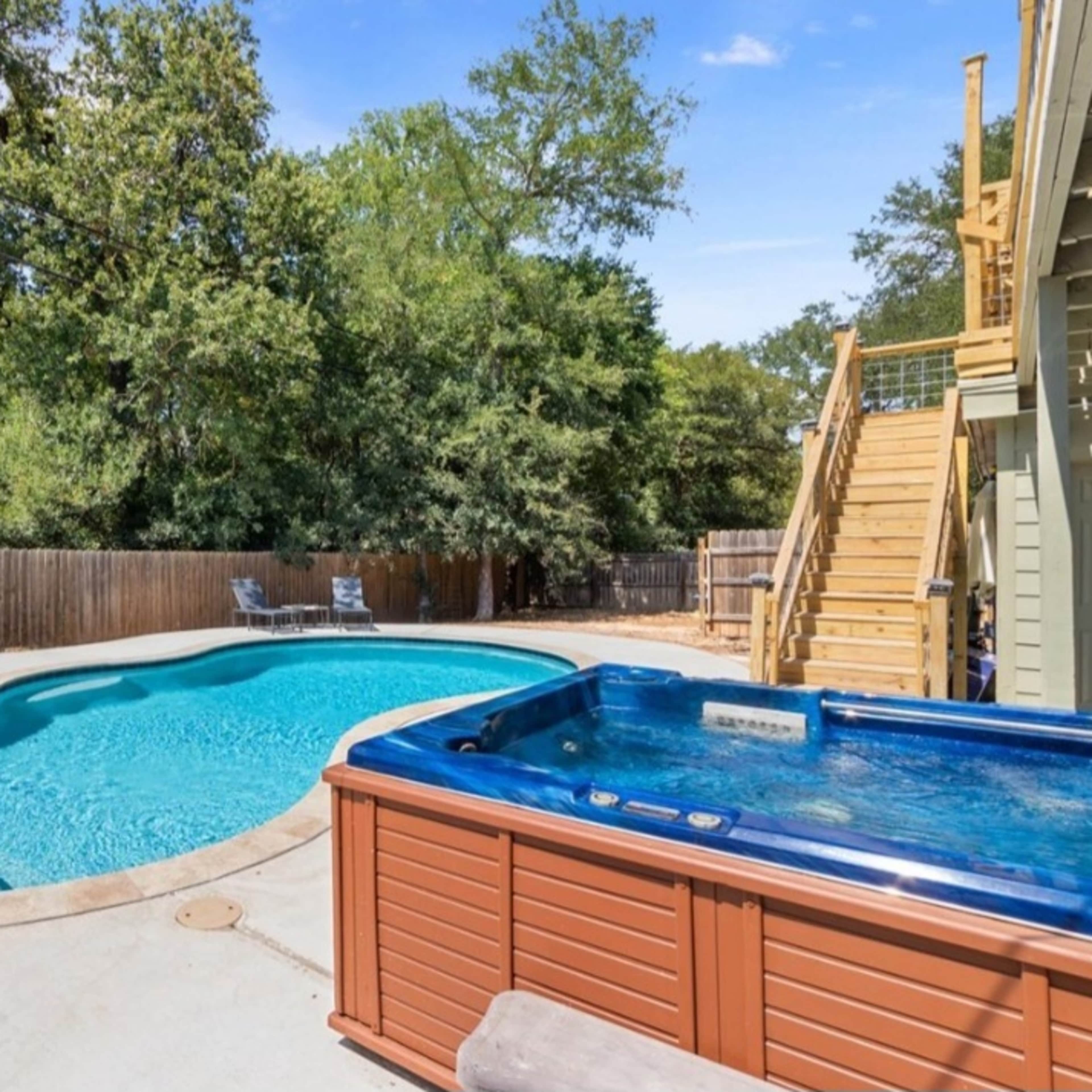 A backyard with a swimming pool and a hot tub, surrounded by trees and a wooden fence, along with lounge chairs nearby.