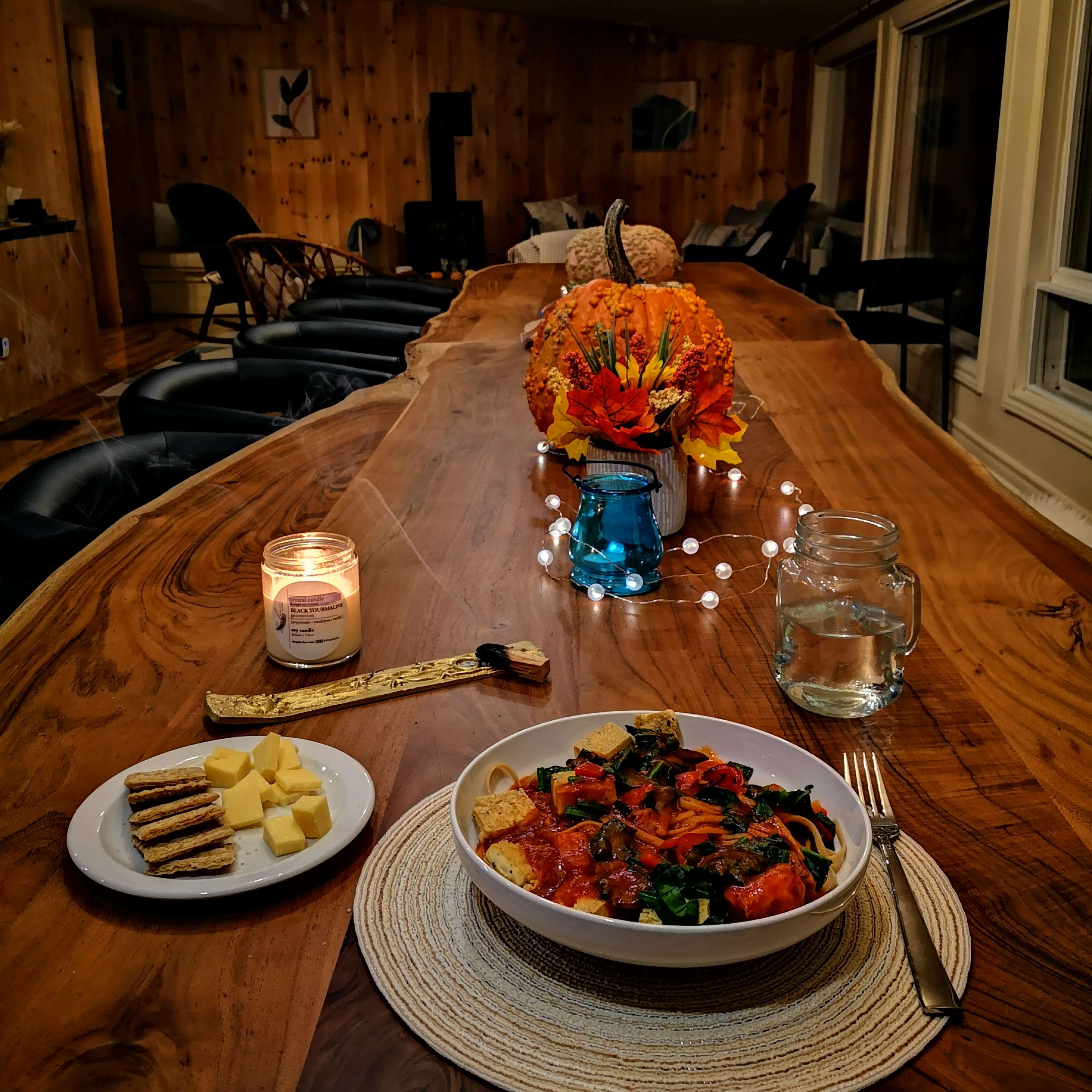 A long wooden table is set for dinner with a plate of pasta, cheese, and crackers, accompanied by a candle and a vase of flowers, in a cozy, warmly lit room.