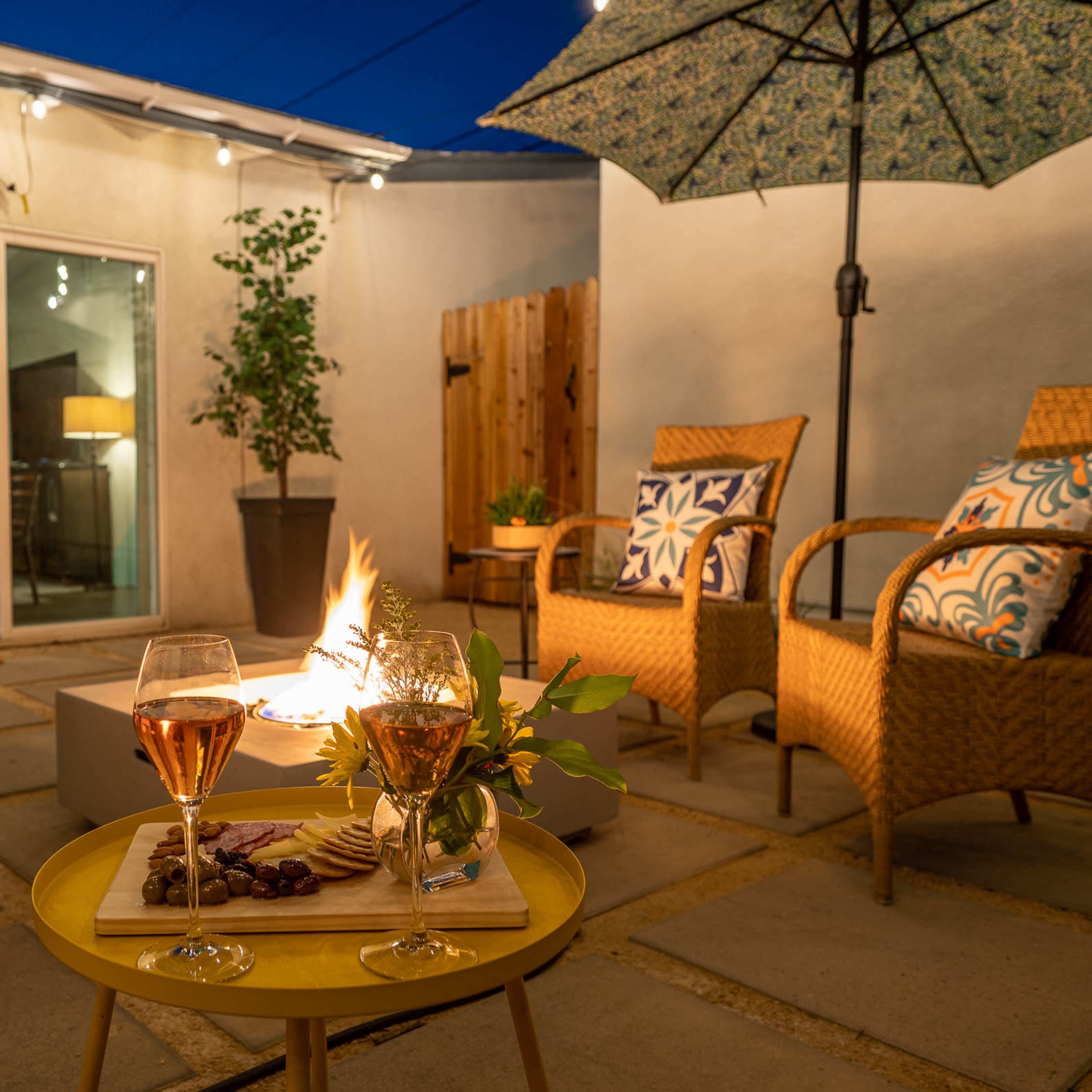 A cozy outdoor seating area features two glasses of rosé wine and a charcuterie board beside a fire pit, surrounded by chairs and umbrellas under a twilight sky.