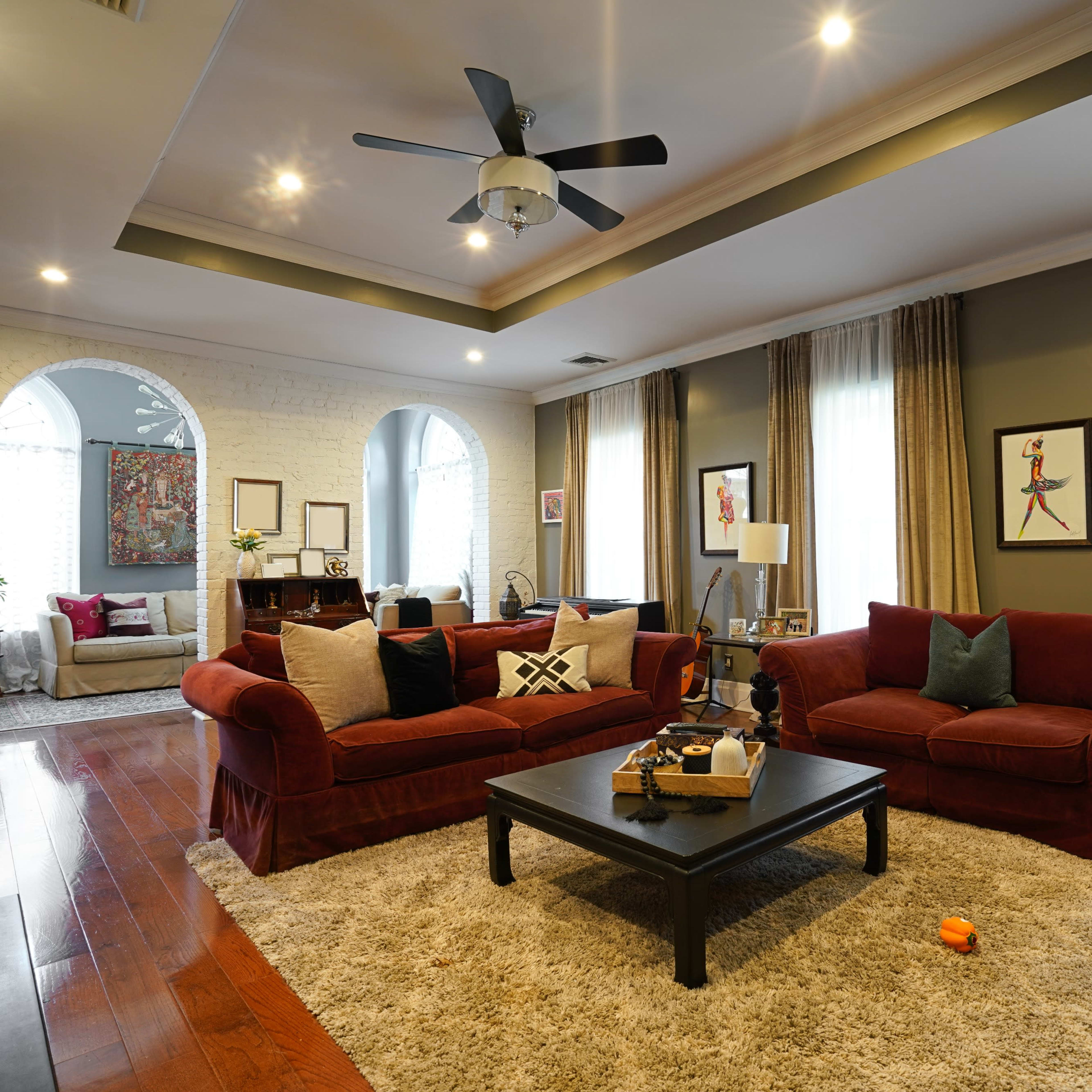 The living room features two red sofas facing a black coffee table, surrounded by large windows with drapes and a soft rug on the wooden floor.