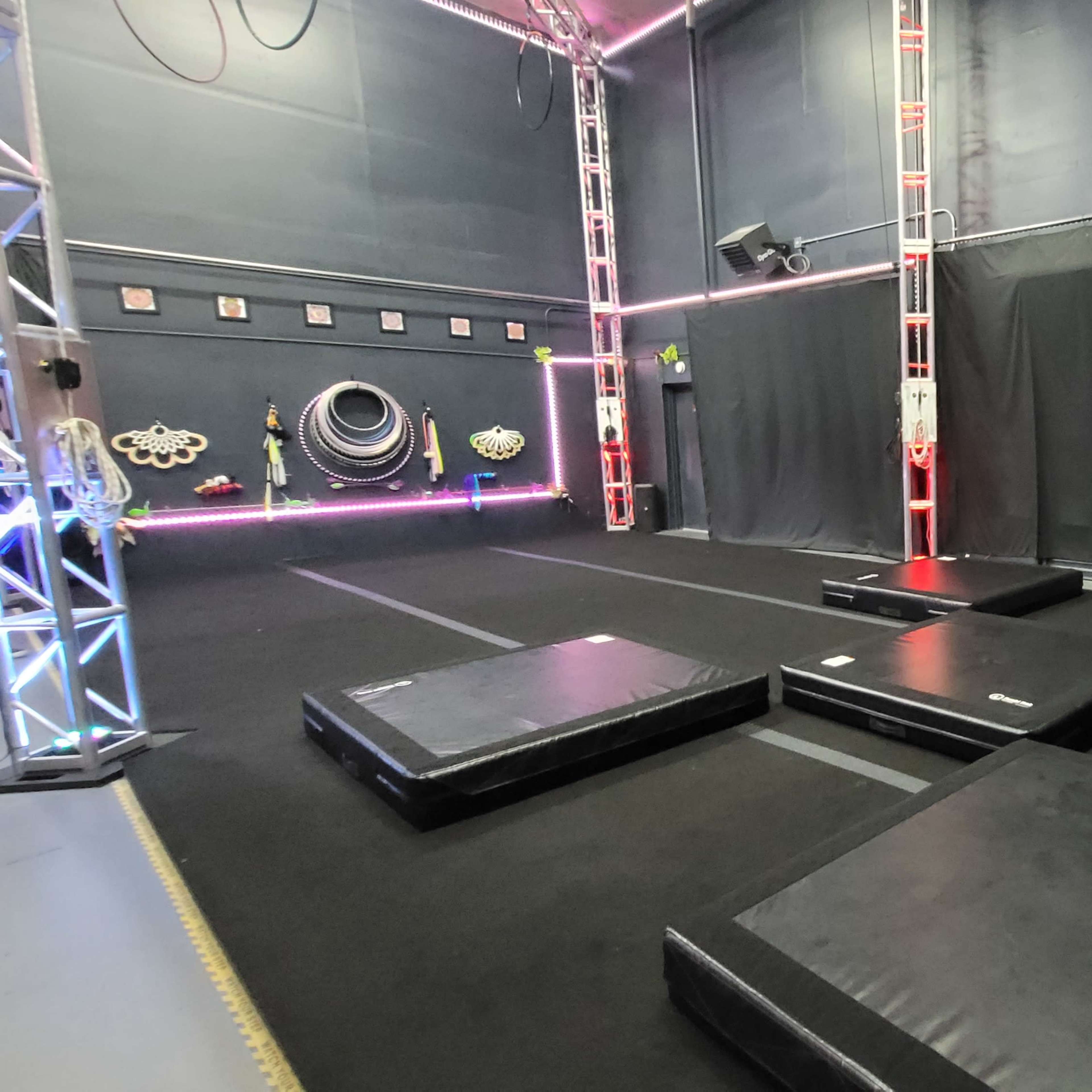 A gymnasium space with several black mats arranged on a dark floor, surrounded by lighting and equipment on the walls.