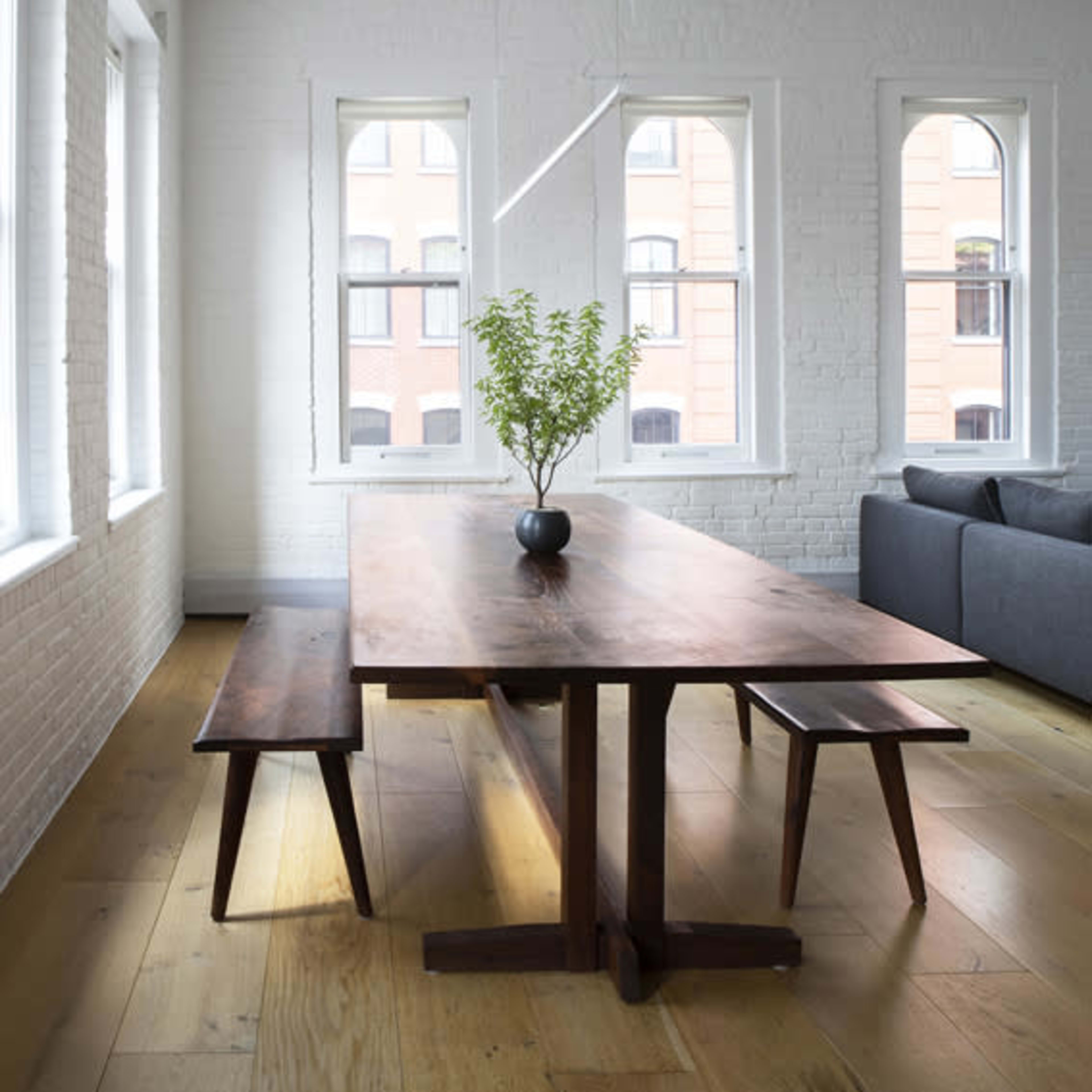 A wooden dining table with a plant centerpiece is positioned between two benches in a bright room with white brick walls and large windows.