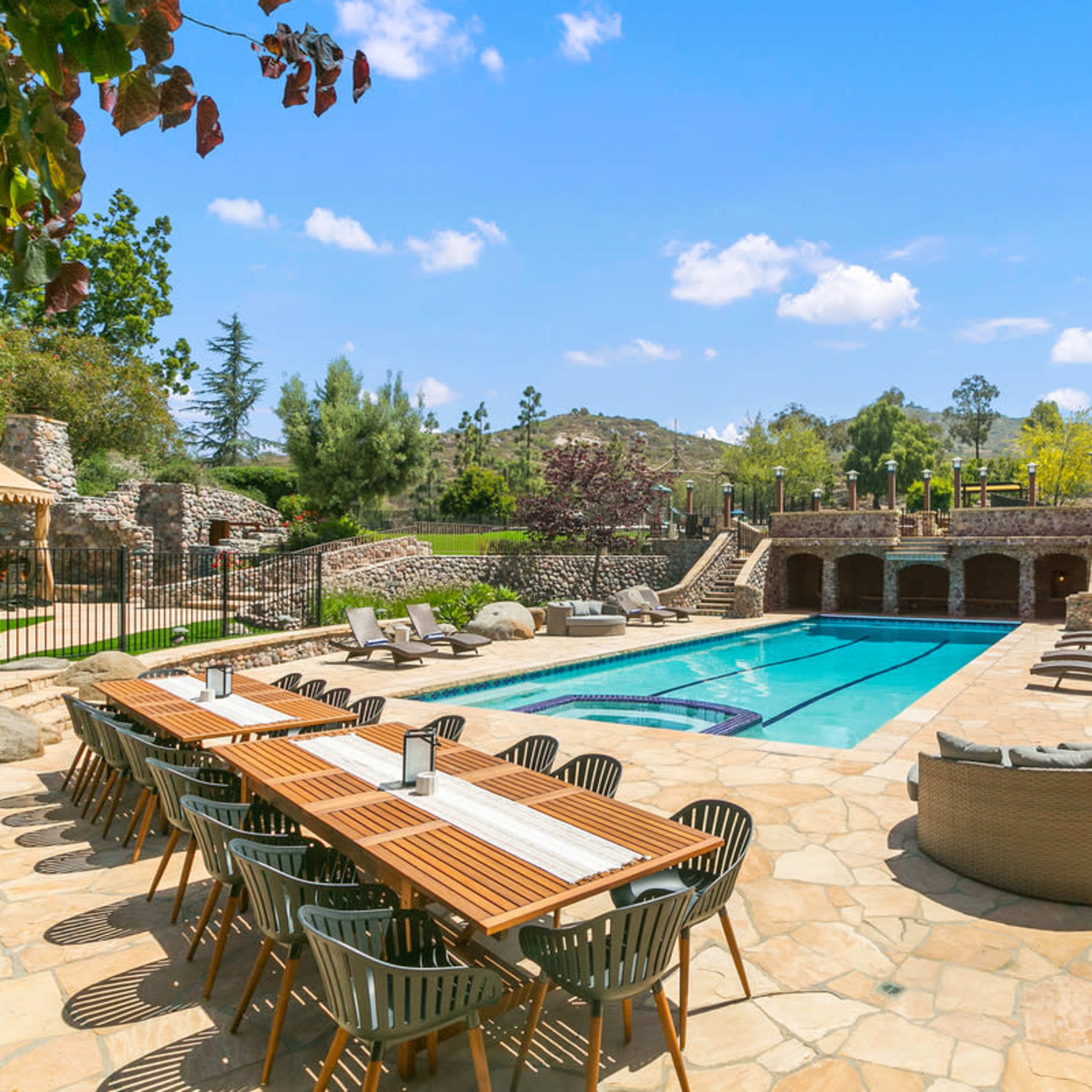 A spacious outdoor area features a swimming pool surrounded by lounge chairs and a long dining table.