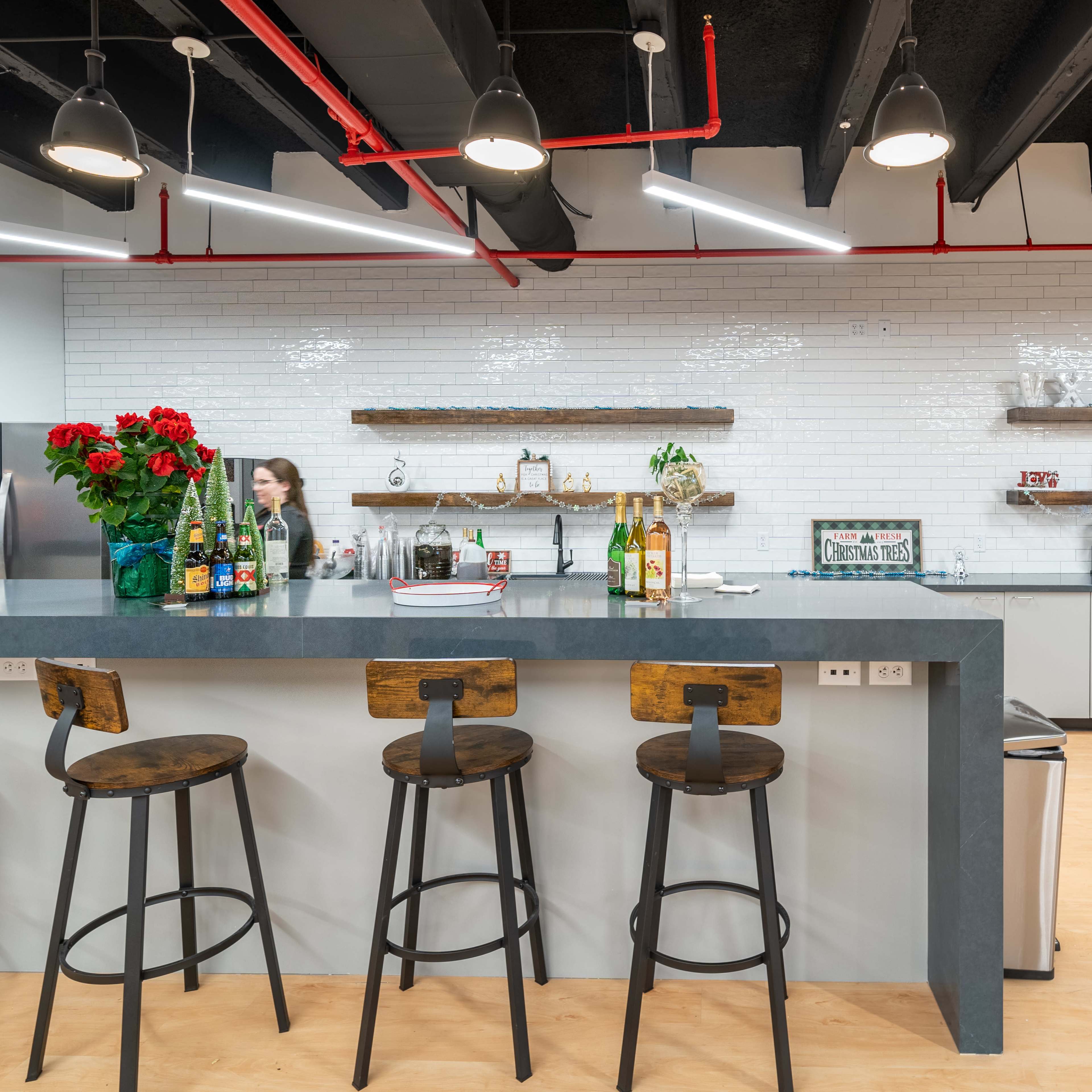 A modern kitchen space features a long gray countertop with high stools, shelves lined with decor, and a variety of beverages displayed.