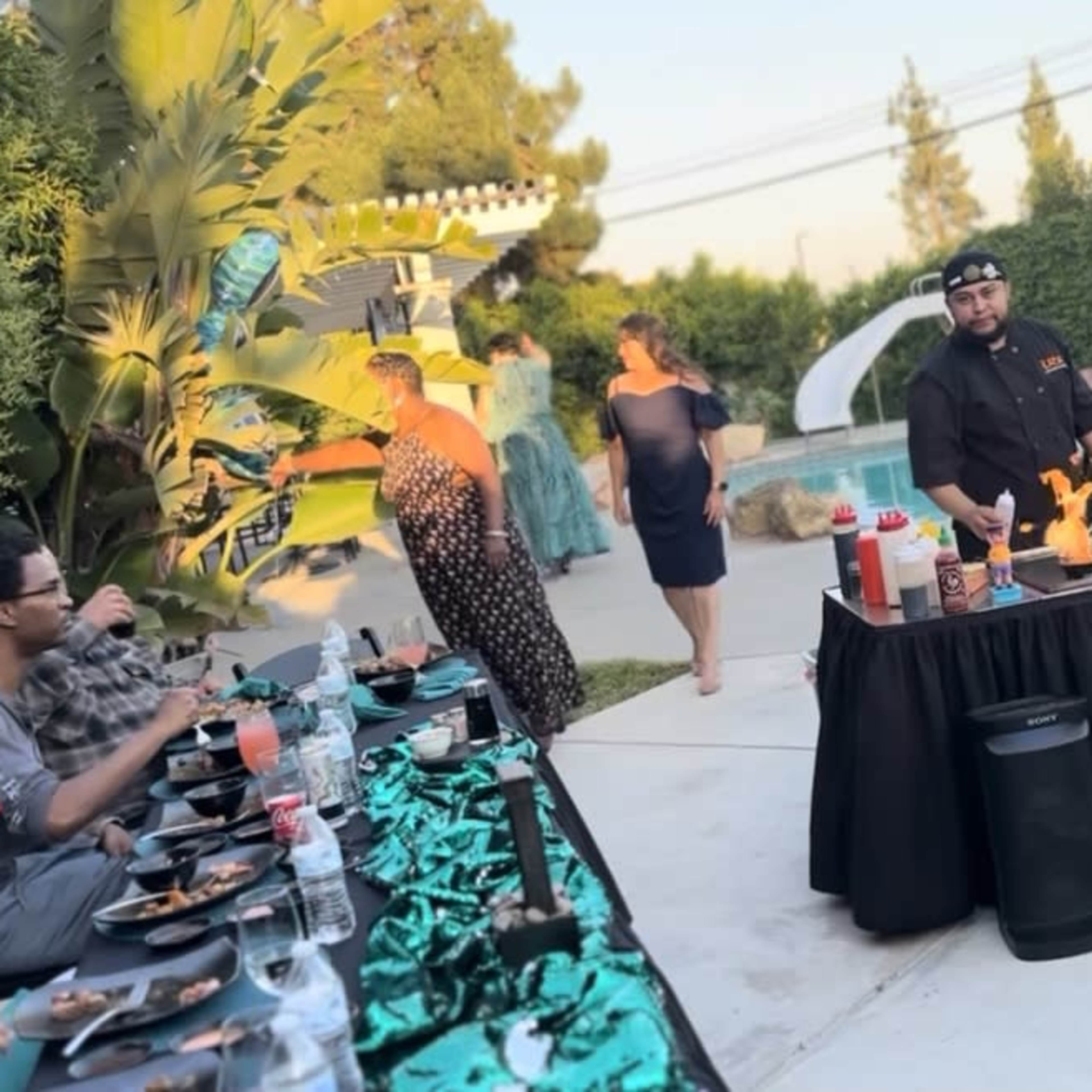 A chef cooks at an outdoor grill surrounded by tables set for a gathering near a swimming pool.