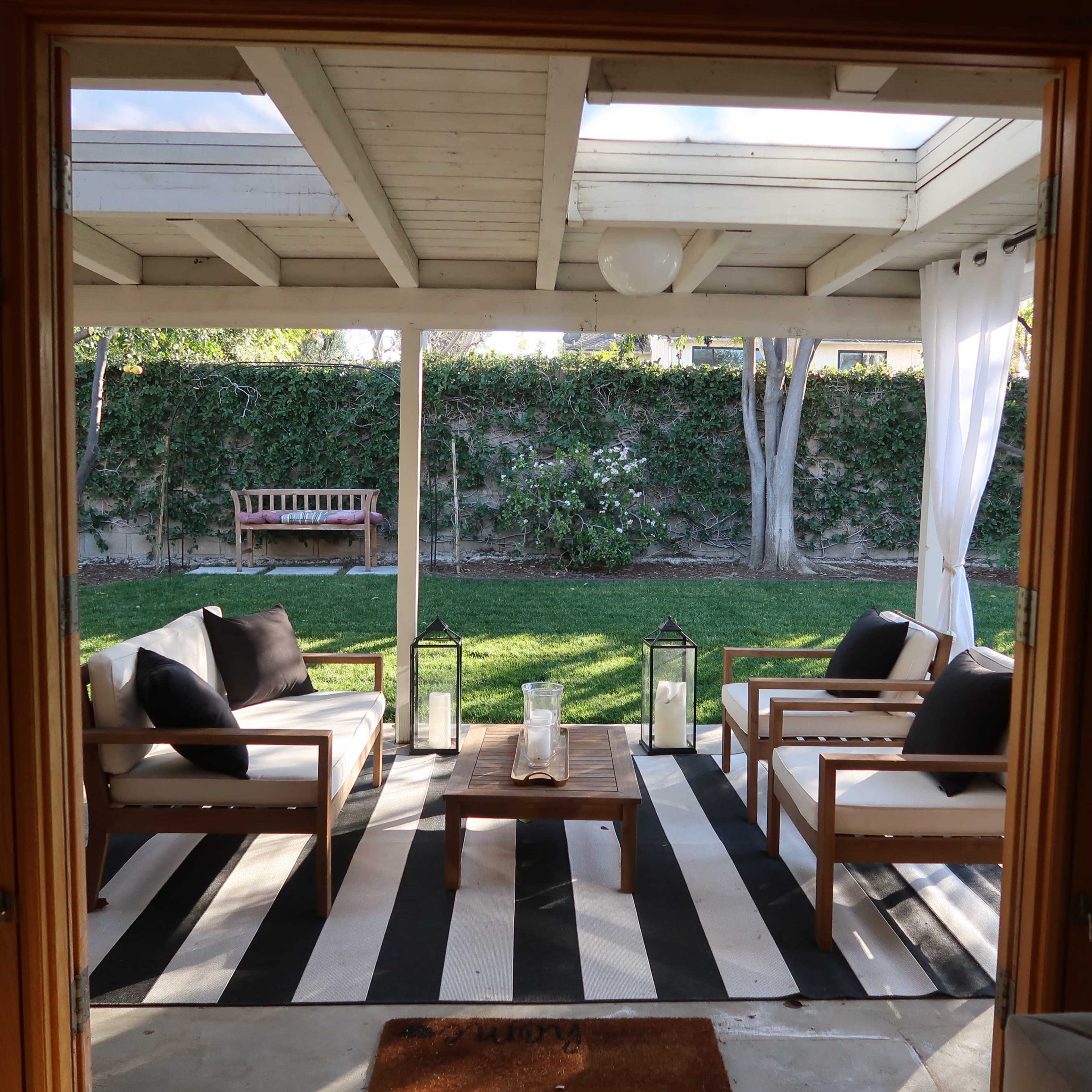 A patio area with wooden chairs, a coffee table on a striped rug, and large windows overlooking a grassy lawn and garden.