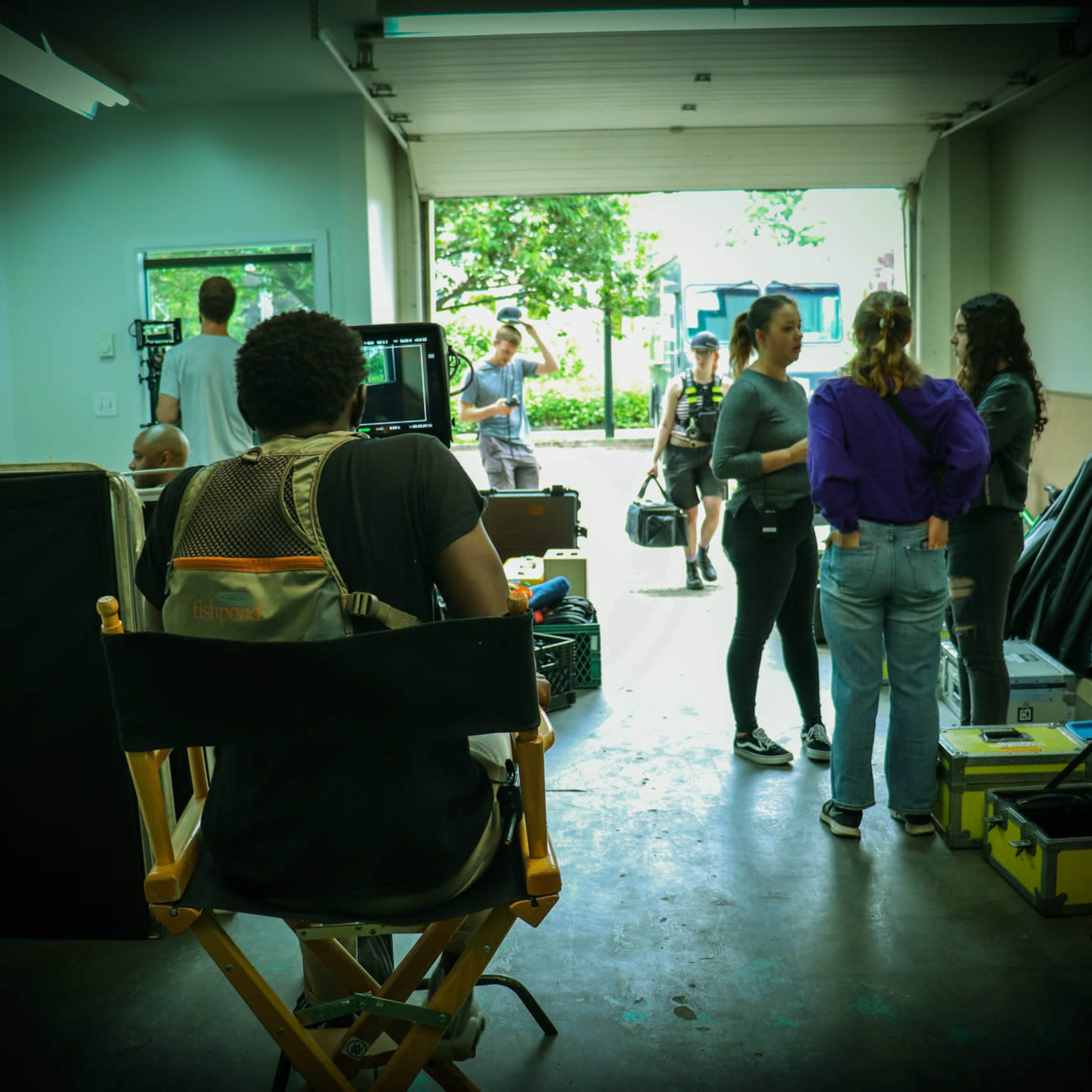 A film crew prepares for a shoot in a garage-like space, with a director seated in a chair facing the action outside.