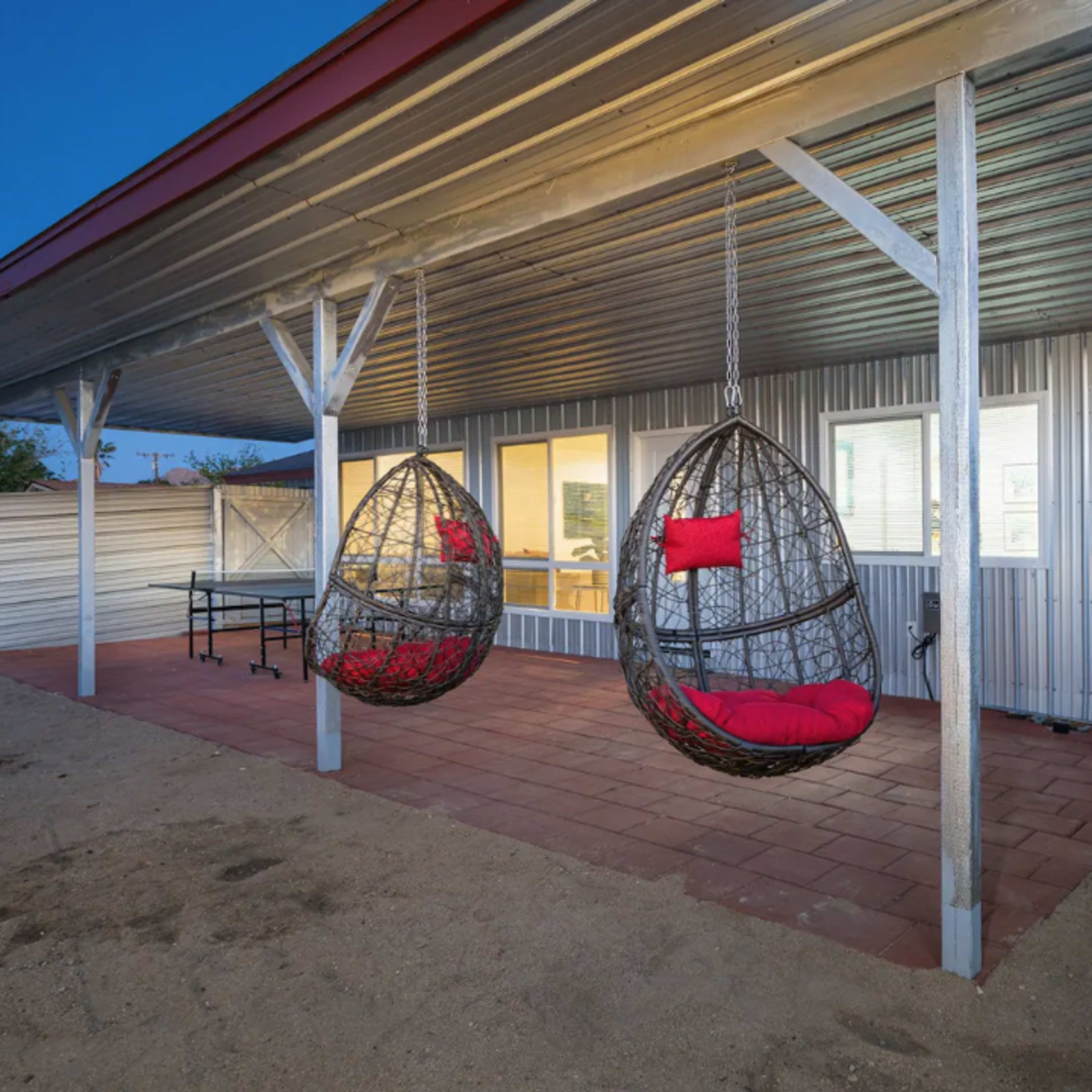 A patio with two hanging chairs and a view of a house with large windows under a metal roof.