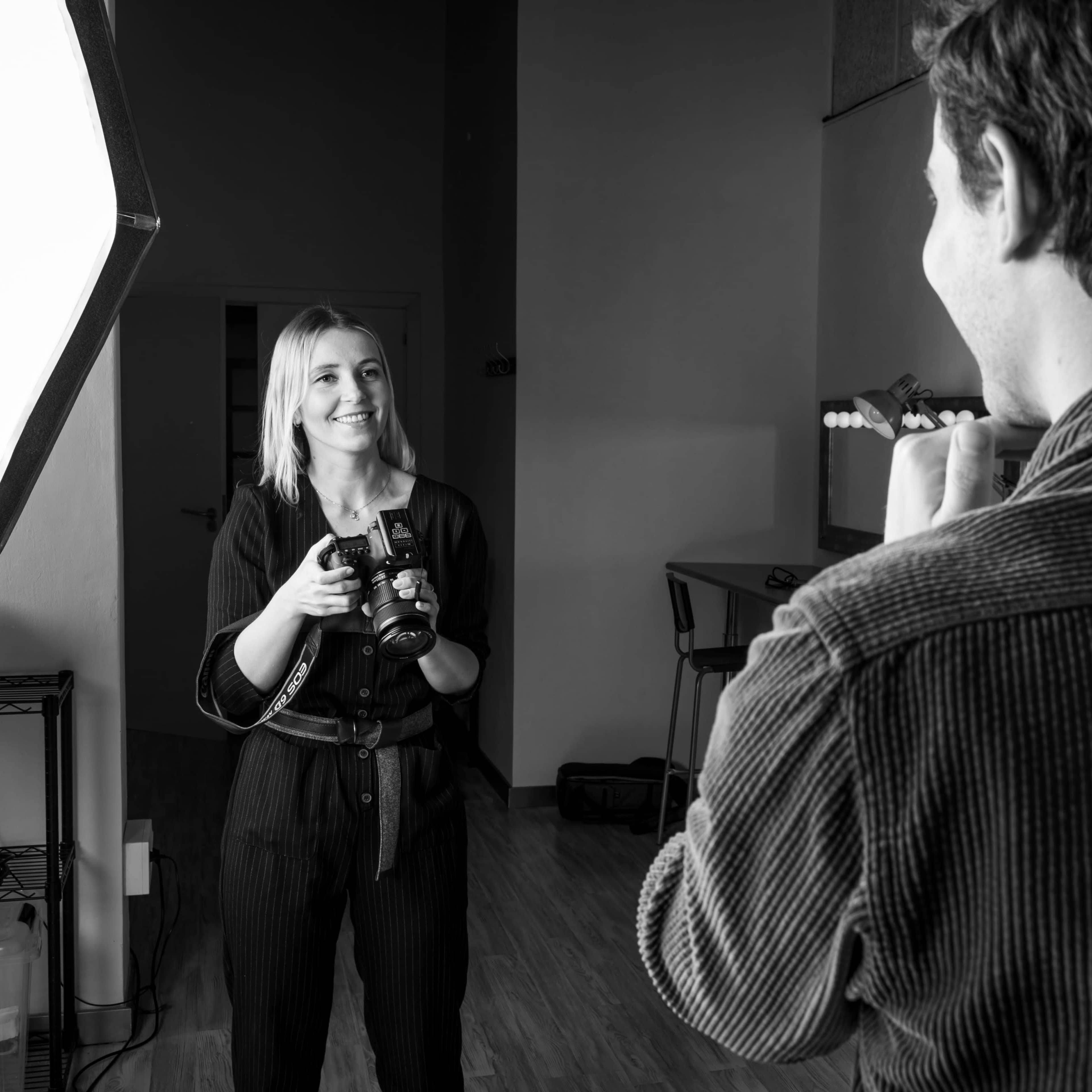 A woman holds a camera while smiling at a man who is adjusting his appearance in front of a mirror.
