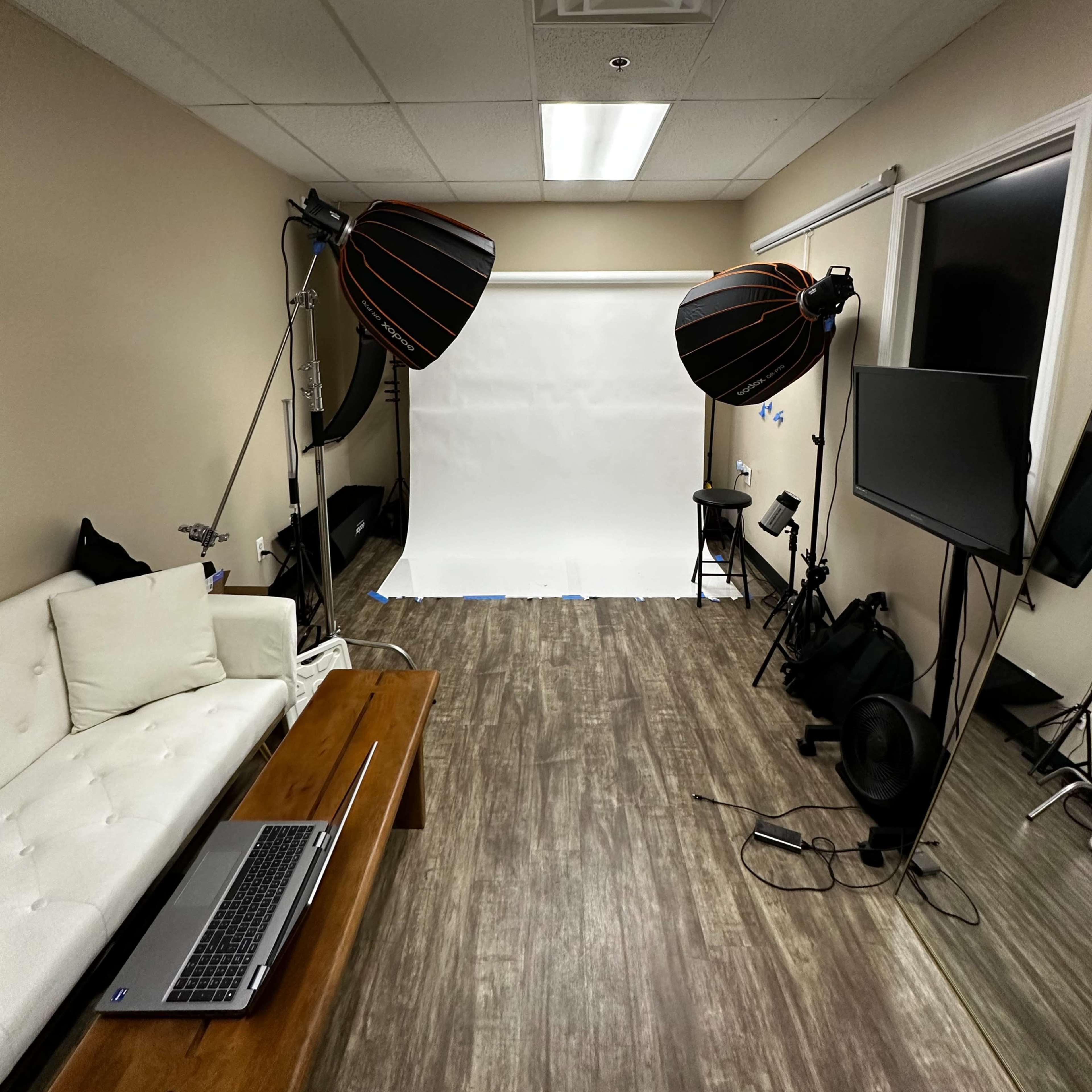 A photography studio with a white backdrop, two large softbox lights, a laptop on a table, and a wall-mounted screen.