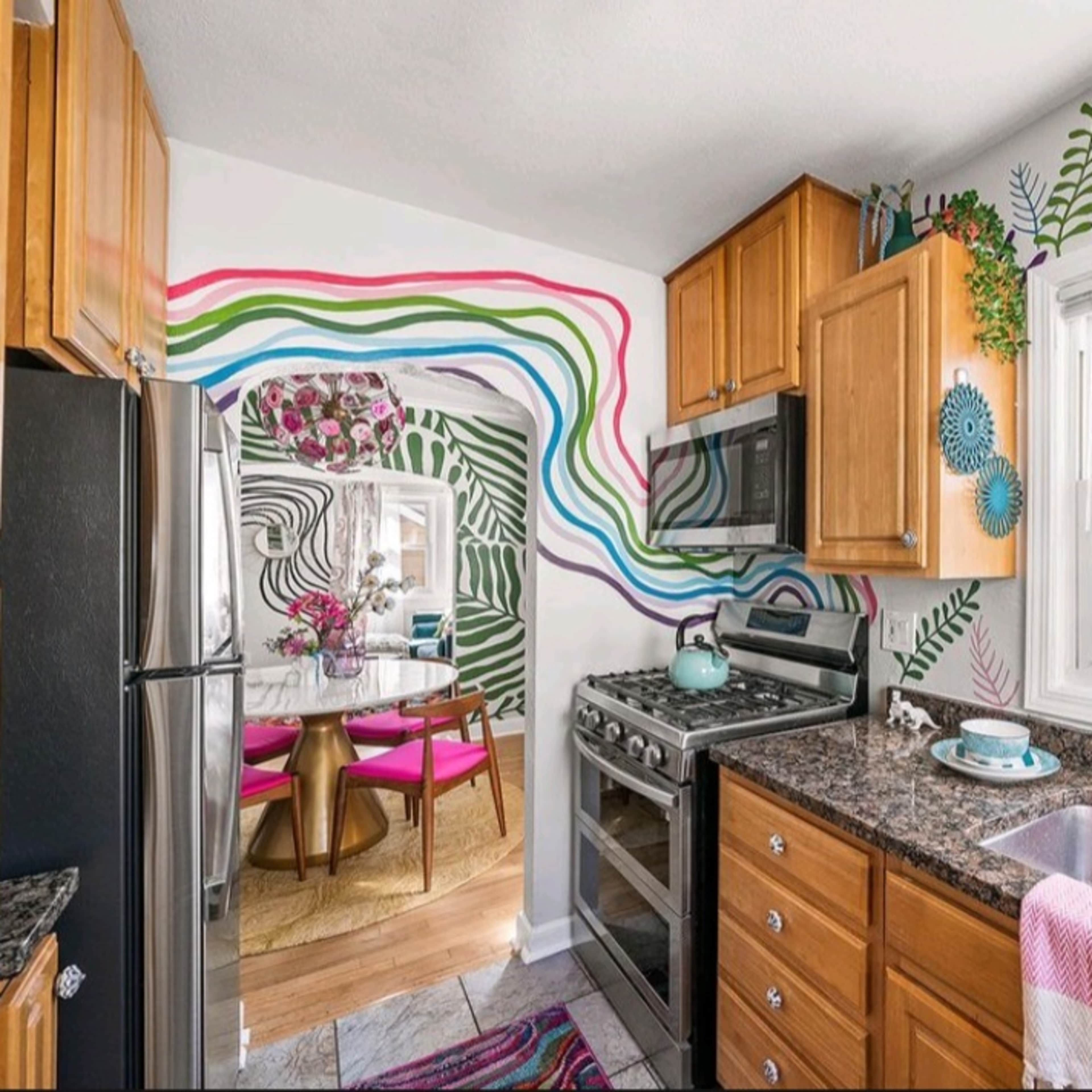 A colorful kitchen features wooden cabinets, a stainless steel stove, and a sink, with vibrant wall art and a view into a dining area with a round table and pink chairs.