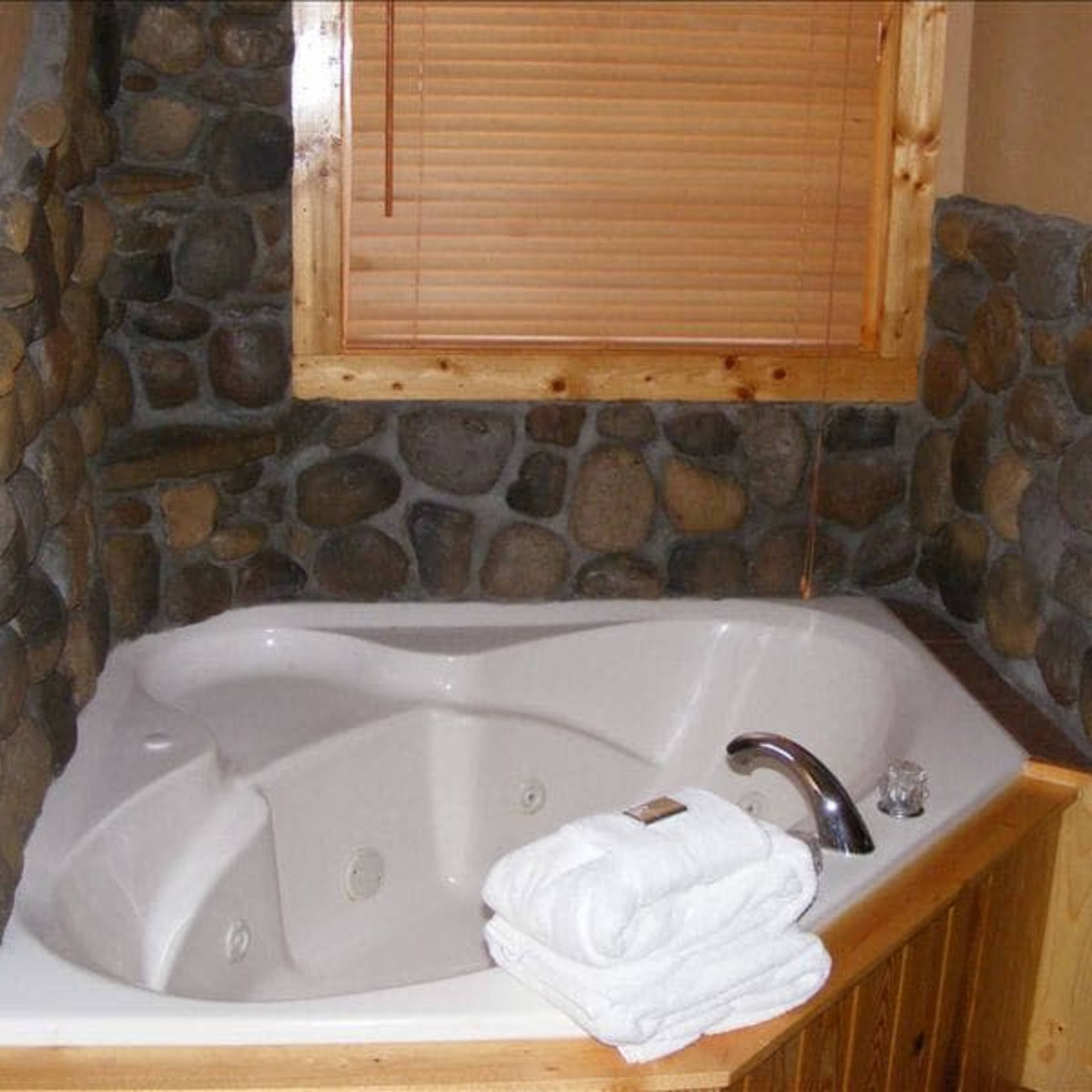 A corner jacuzzi tub is surrounded by a stone wall and wooden accents, with neatly folded white towels placed on the edge.