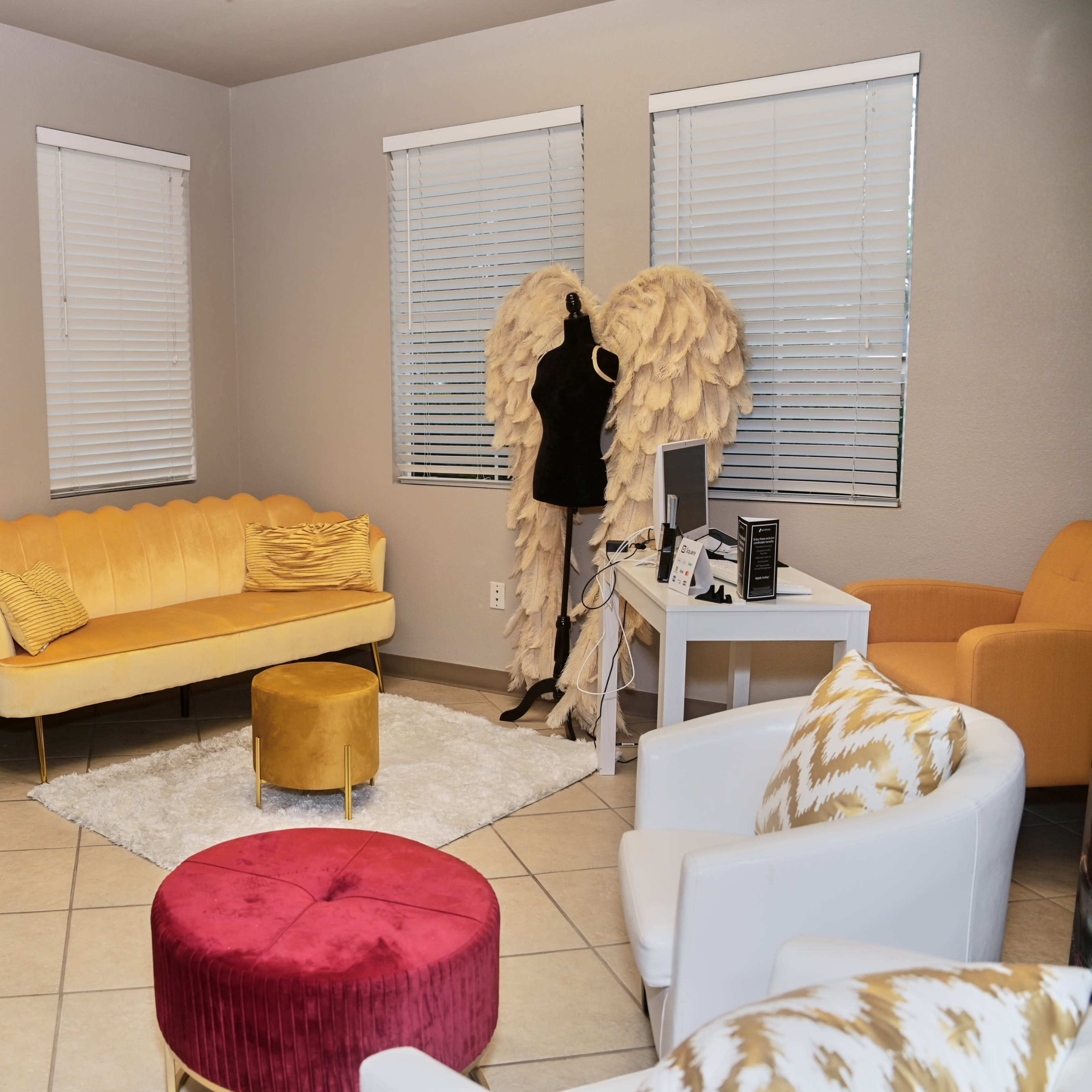 A stylish waiting area with a yellow couch, a round ottoman, a mannequin with angel wings, and a desk against a light gray wall.