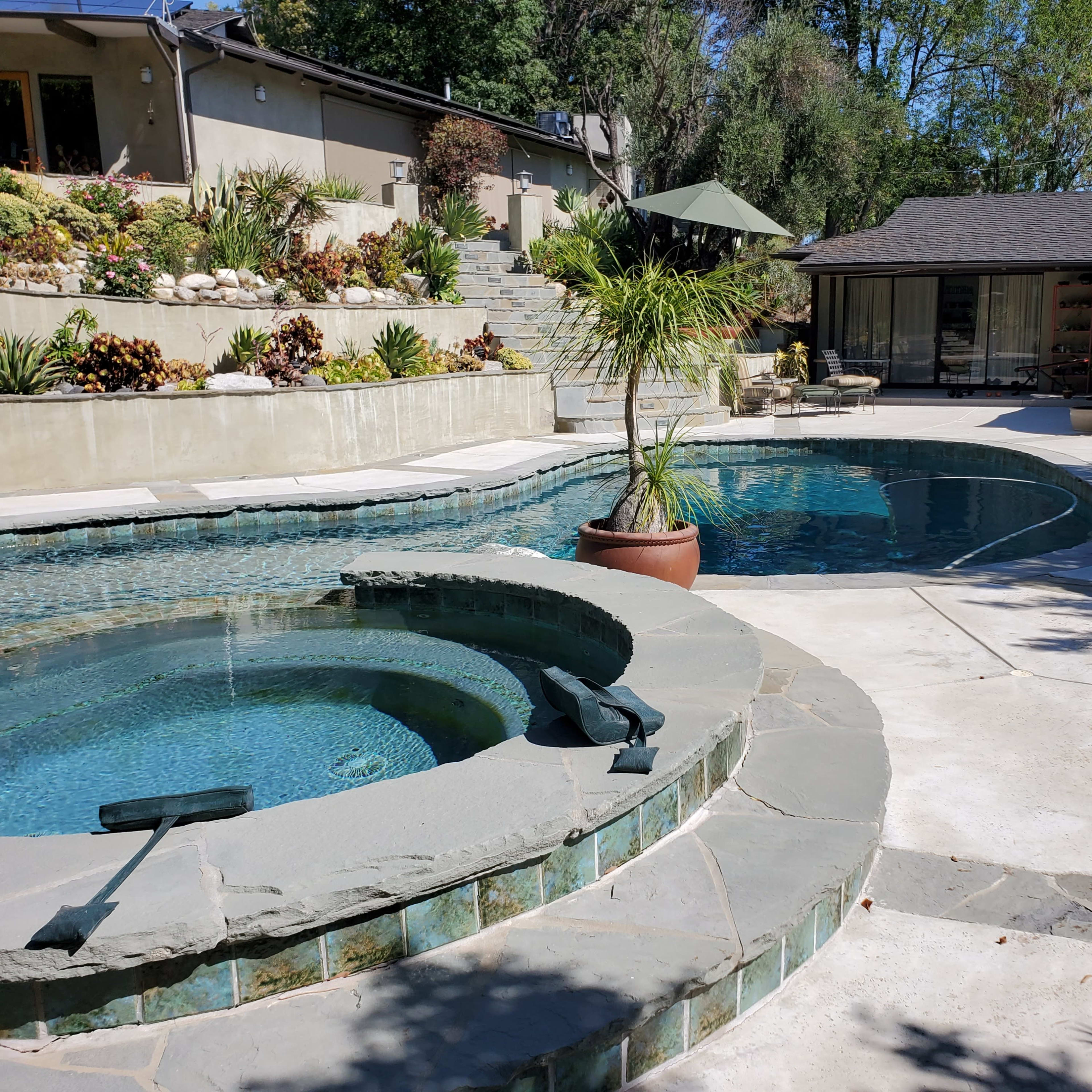 A landscaped pool area featuring a hot tub, a larger swimming pool, and surrounding greenery with a patio.