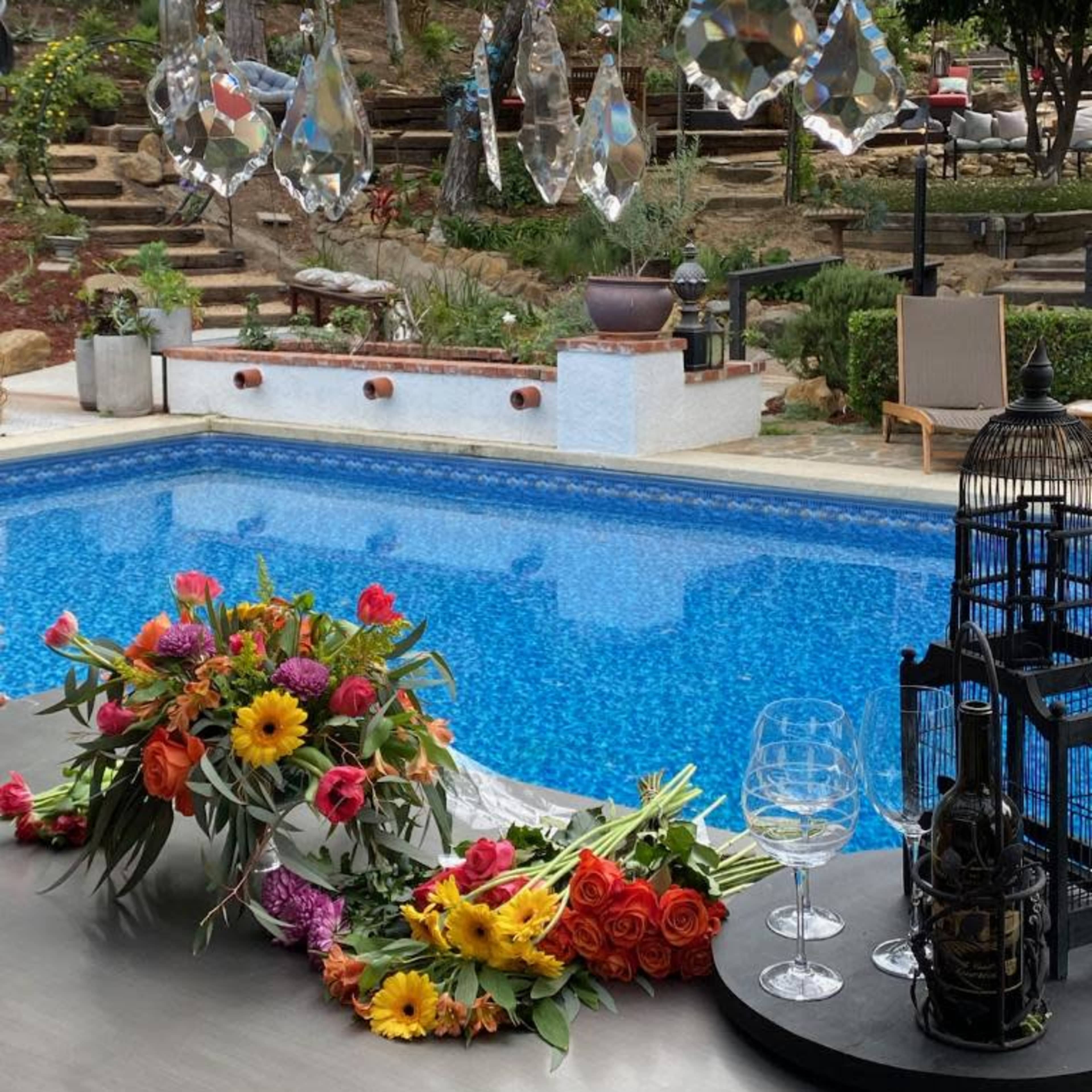 A brightly lit pool area features a blue swimming pool, surrounded by flowers and a decorative chandelier overhead.
