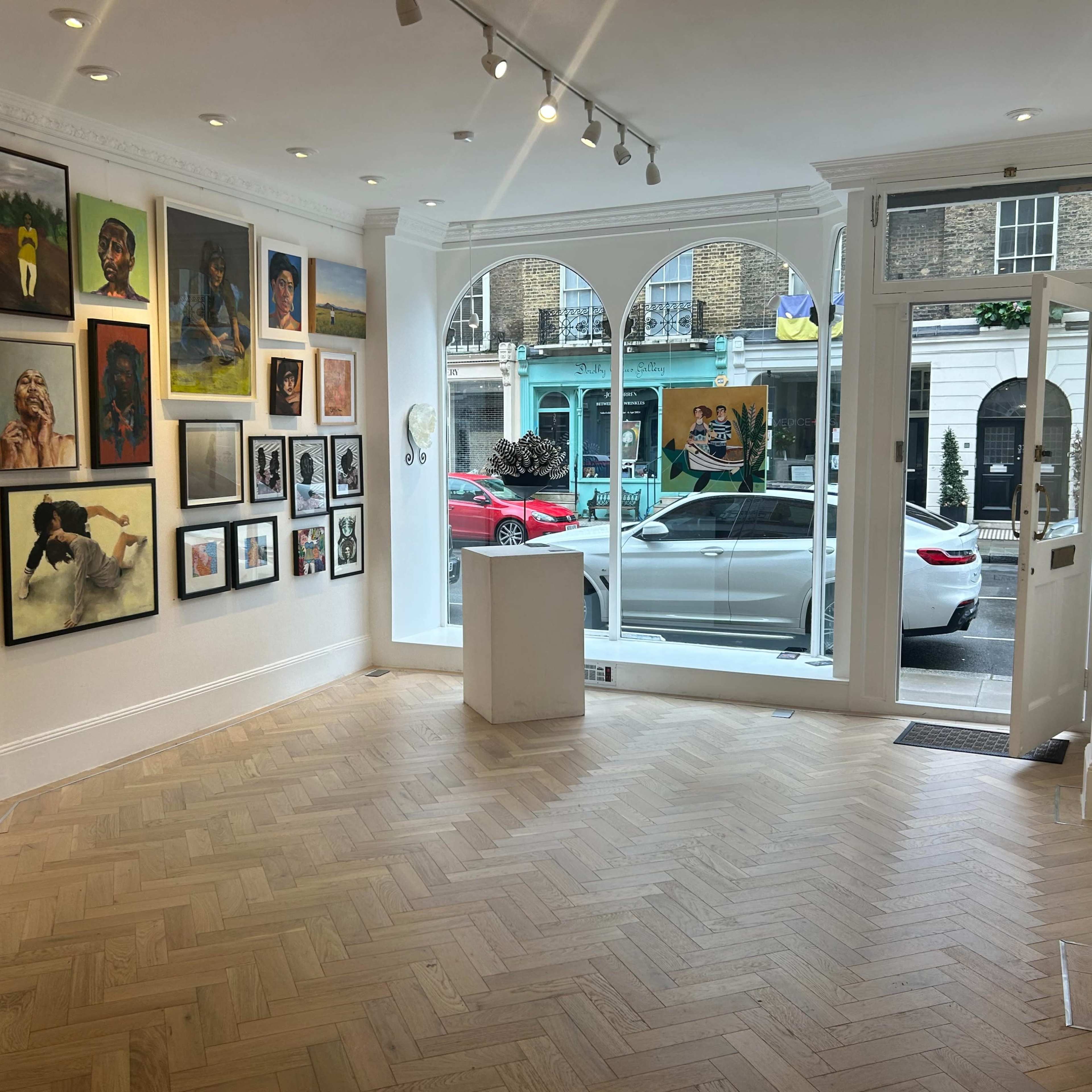 An art gallery with various framed artworks on the white walls and a large window displaying an outdoor street scene with parked cars.