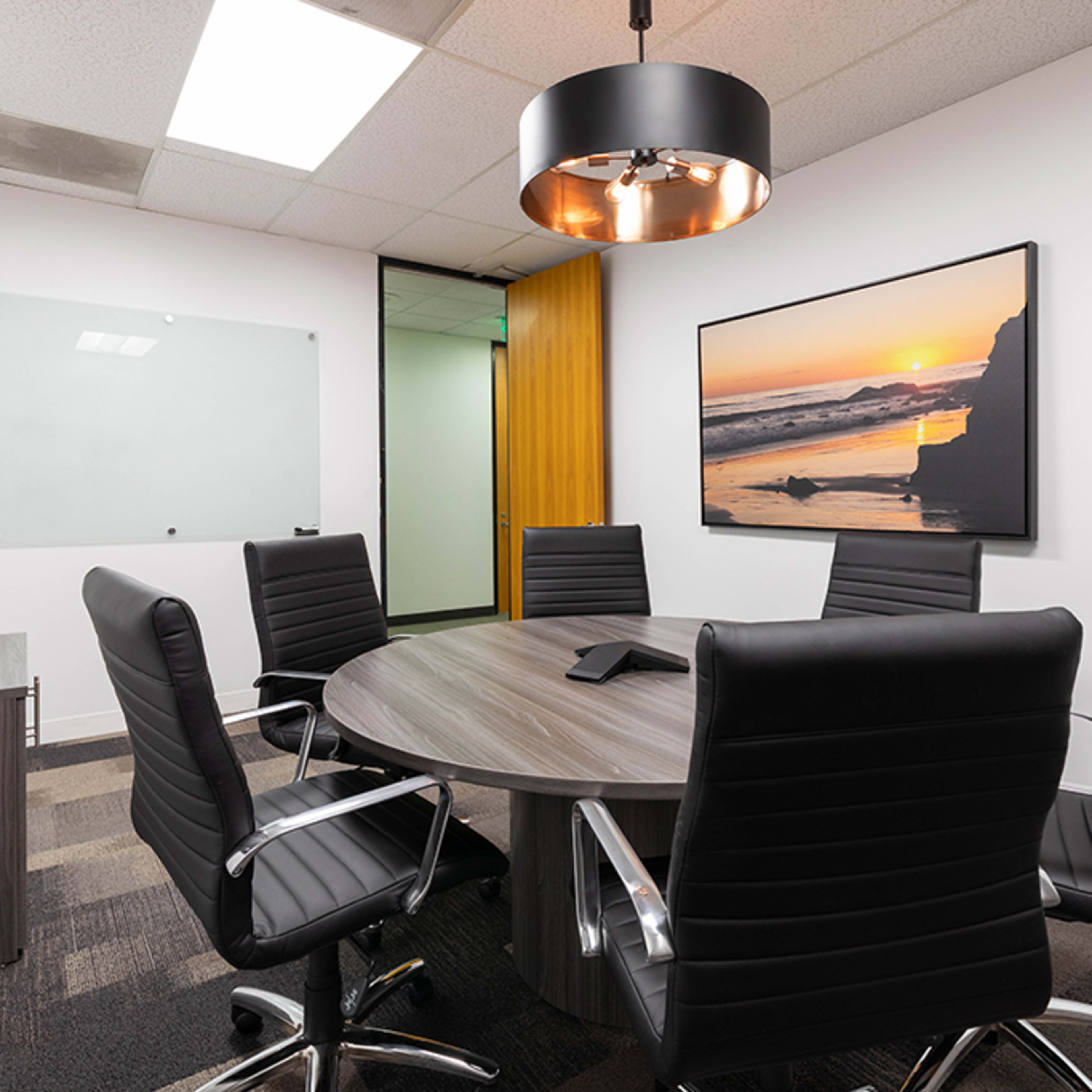 A conference room features a circular table surrounded by black leather chairs, with a large wall-mounted picture of a sunset and a glass whiteboard nearby.