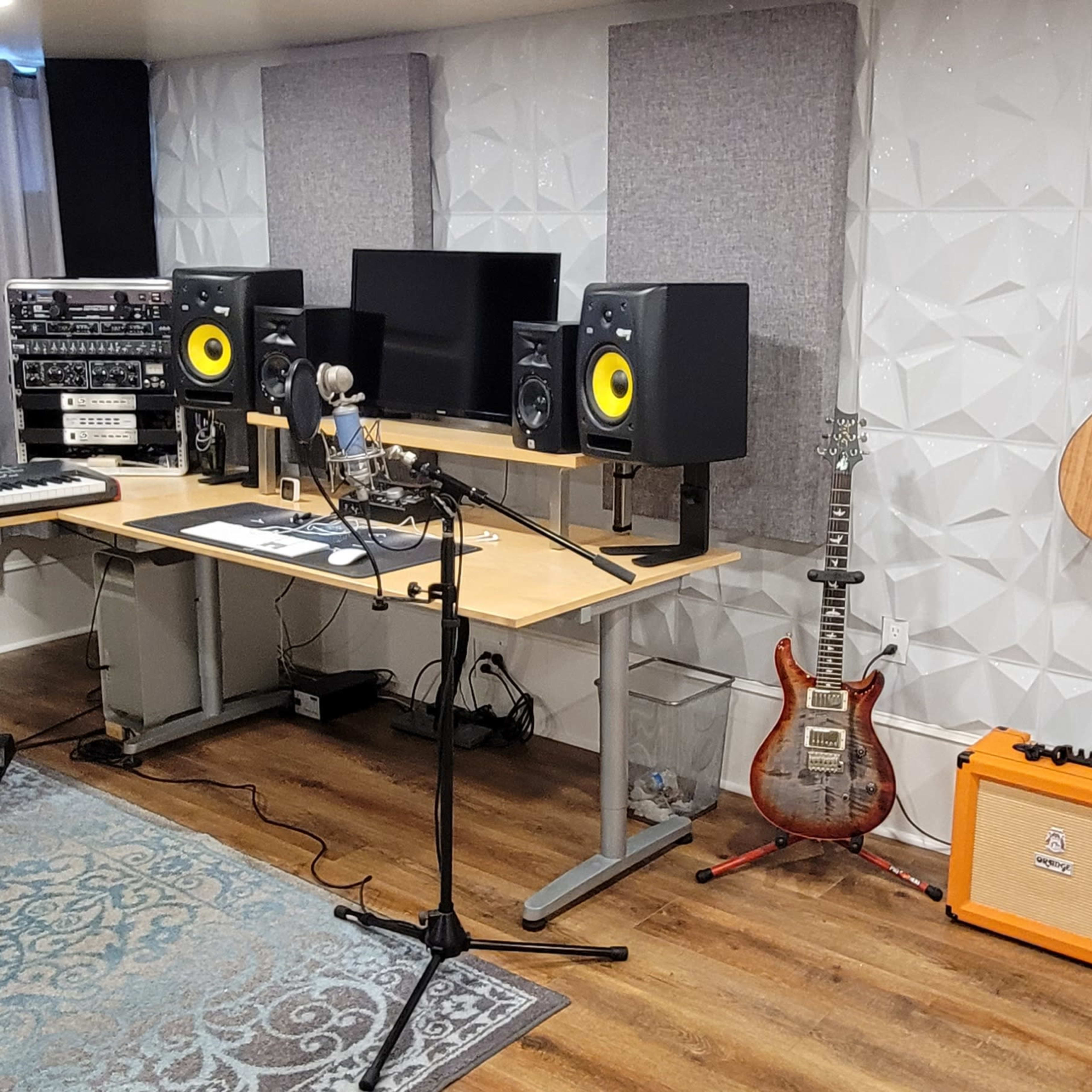 A music recording studio with various instruments, including acoustic guitars, a keyboard, studio monitors, and audio equipment, arranged around a desk with a microphone and chair.