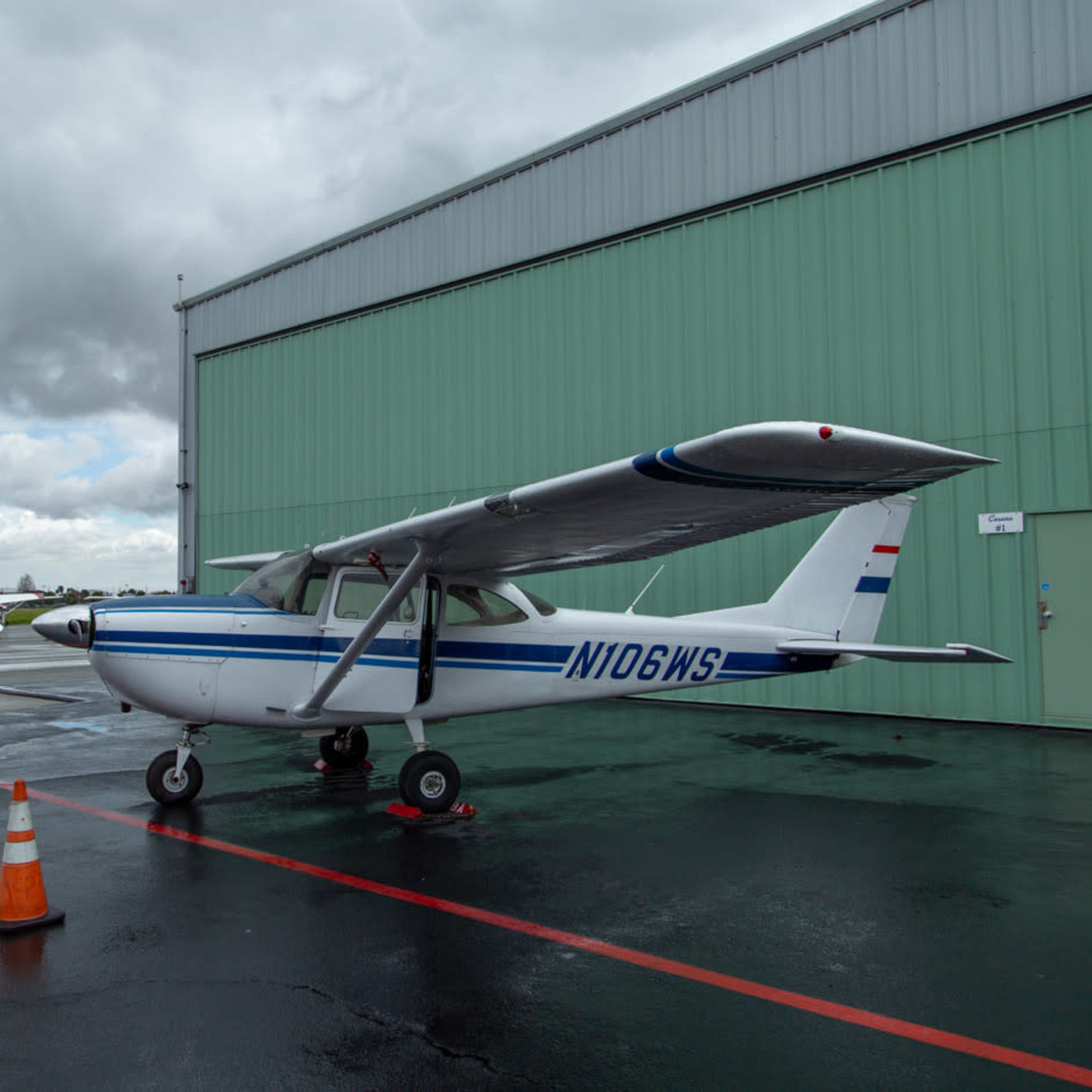 A single-engine Cessna aircraft is parked on a tarmac in front of a green hangar, with orange traffic cones marking its position.