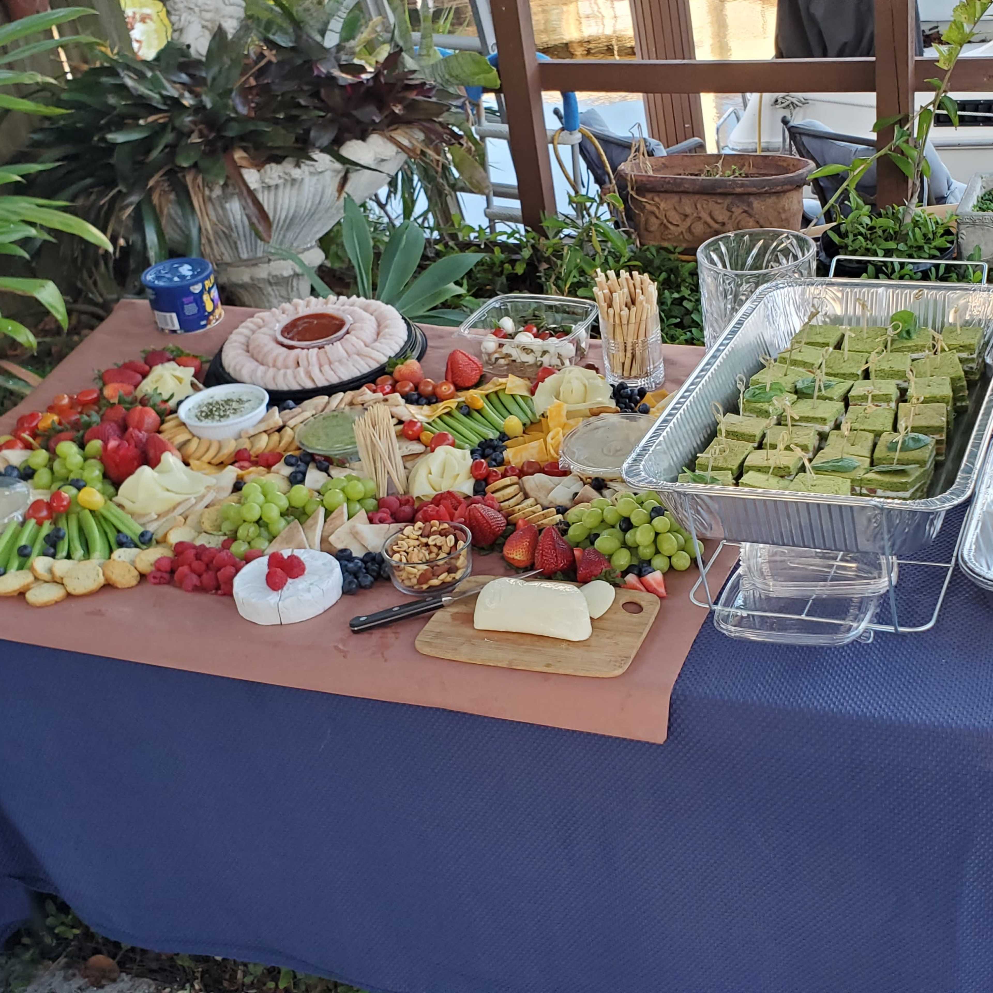 A buffet table is arranged with a variety of food, including fruits, vegetables, cheese, crackers, and desserts, set in an outdoor space.
