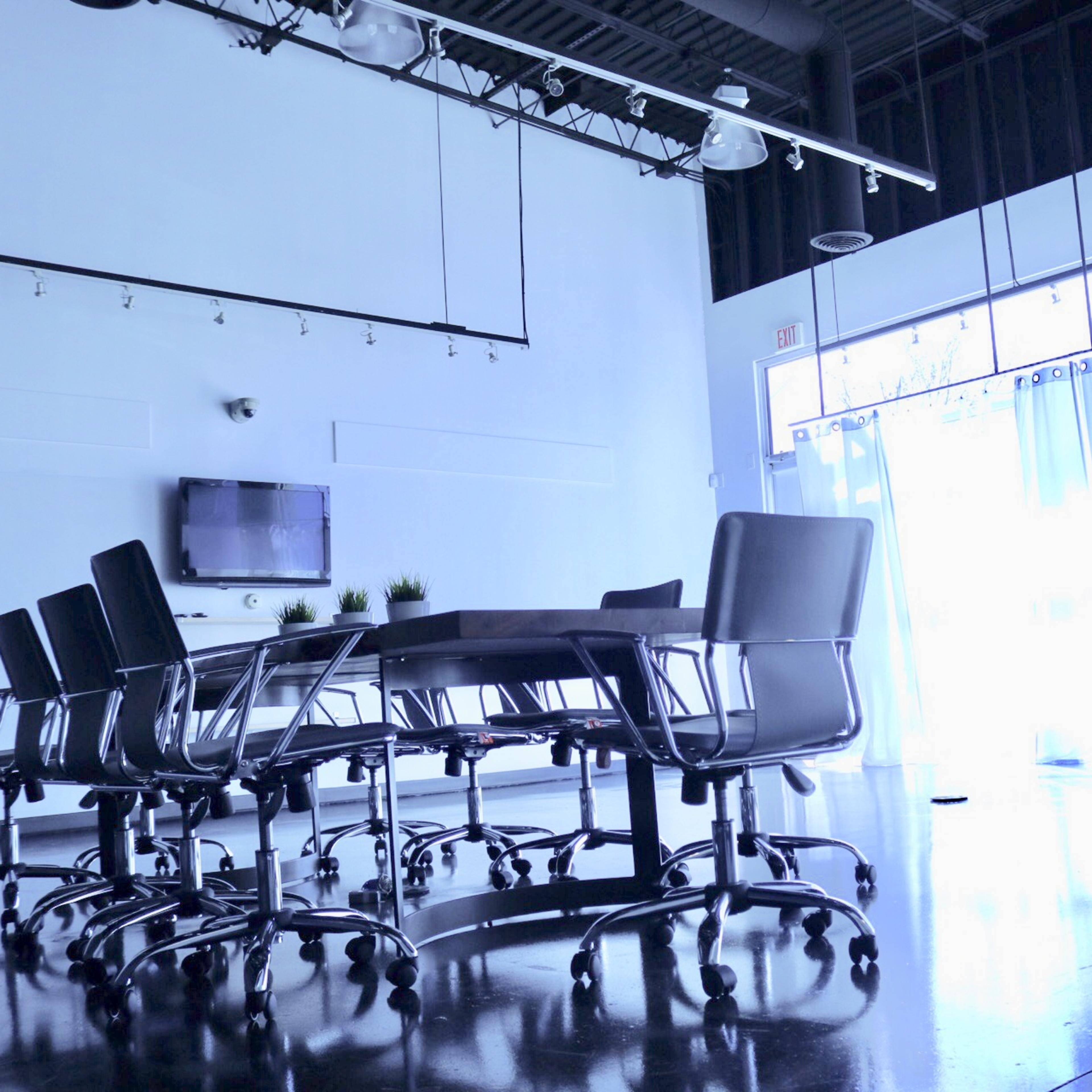 A modern conference room features a large table surrounded by black rolling chairs, with large windows and white curtains on one side.