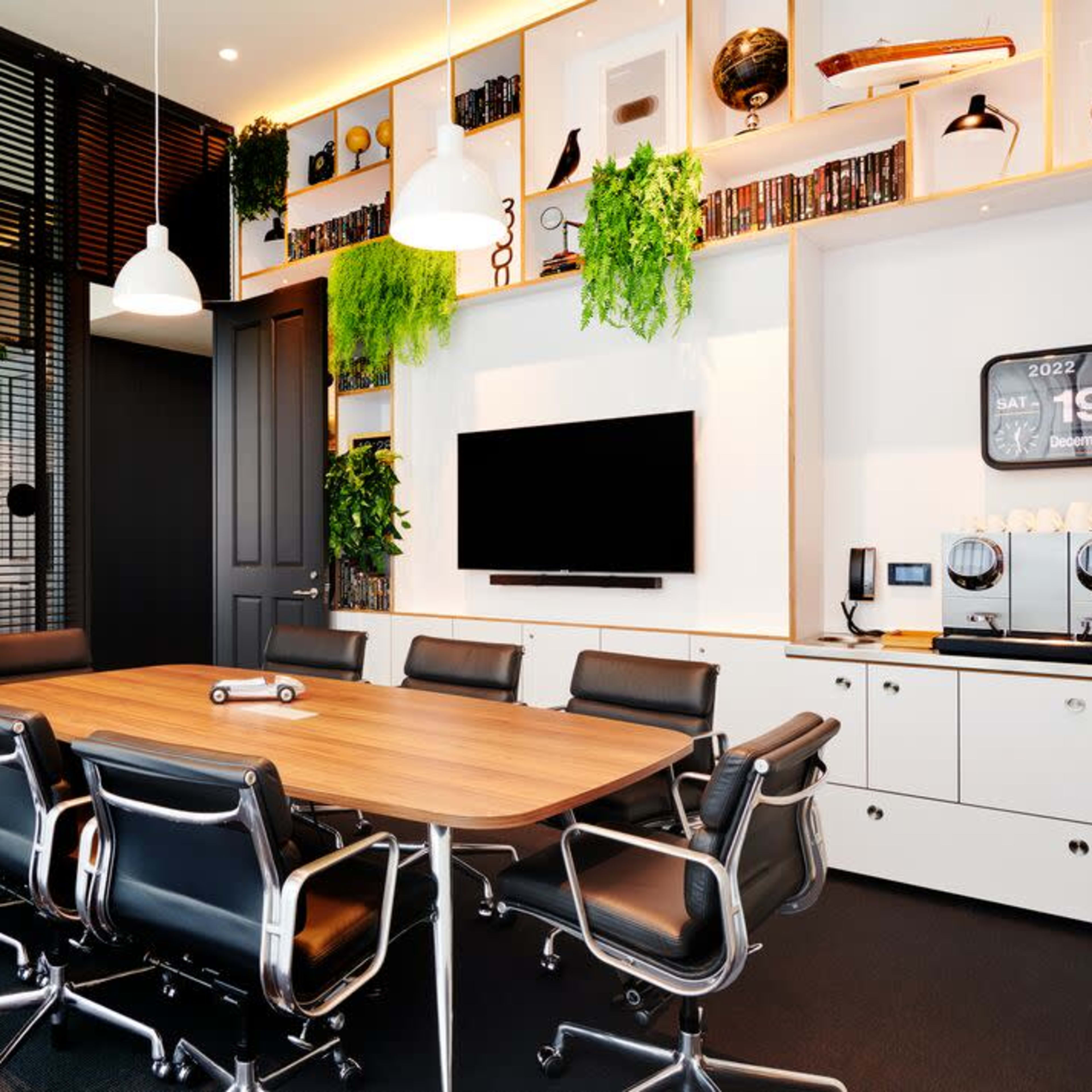 A modern conference room features a large wooden table surrounded by black chairs, with greenery on the walls and a television mounted across from the table.