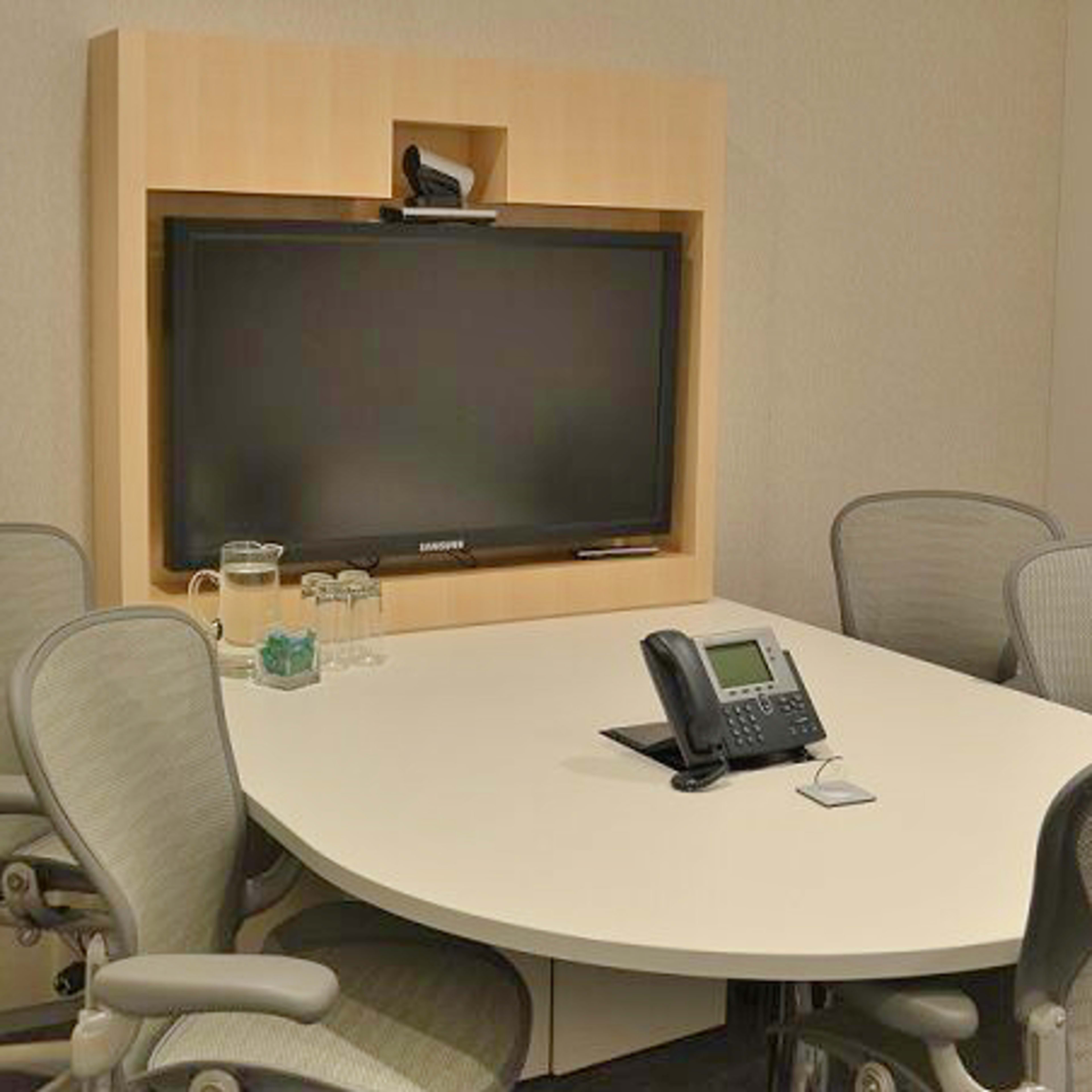 A small conference room with a large wall-mounted television, a round table, and four ergonomic chairs.